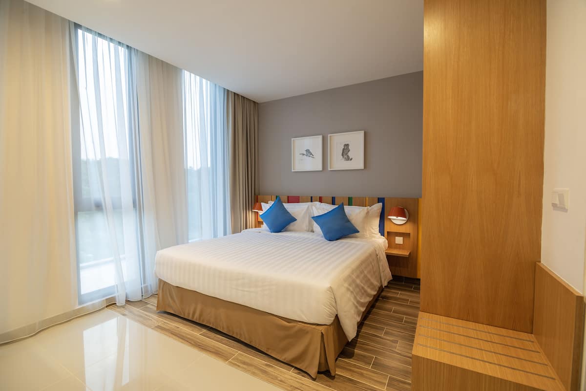 Ein Hotelzimmer mit einem großen Bett, einem Holz-Kopfteil, zwei gerahmten Bildern an der Wand und einem großen Fenster mit durchsichtigen Vorhängen.