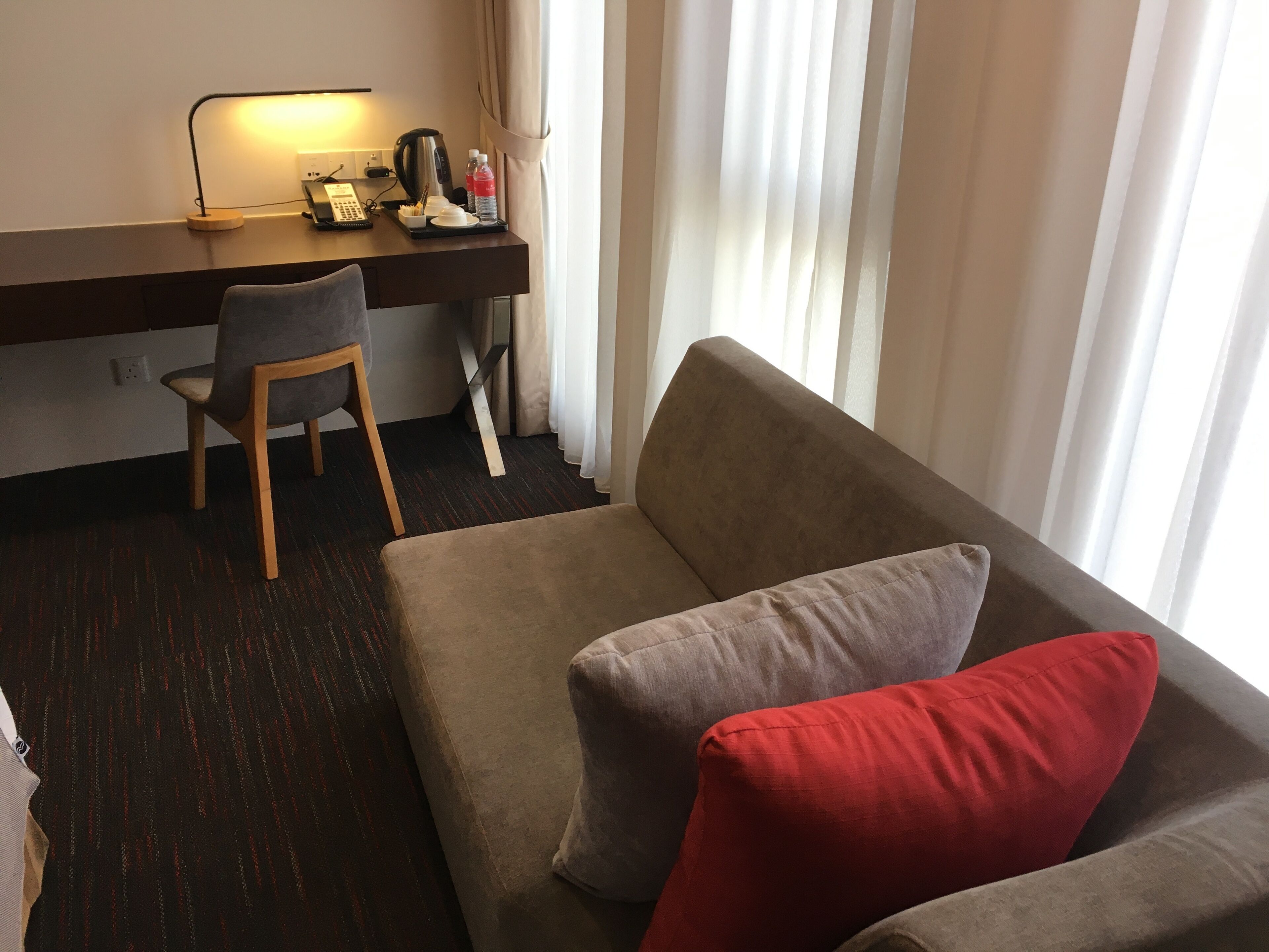 Habitación de hotel con sofá, escritorio con lámpara, una silla y ventana con cortinas.