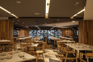 A spacious dining area with multiple tables and chairs, wooden paneling on the walls, and large windows allowing natural light.
