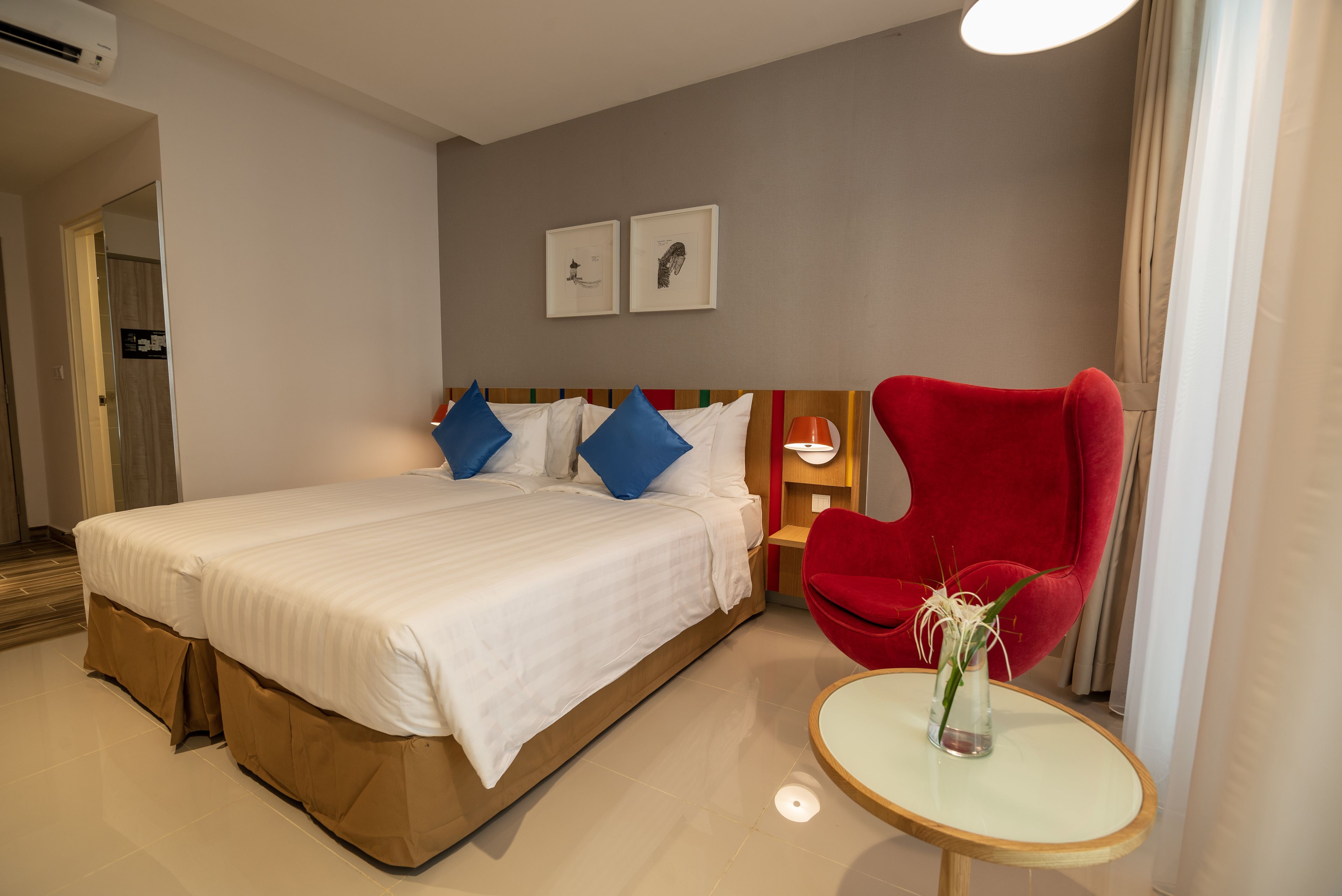A hotel room with a large bed, a red armchair, a small table with a vase, and two framed pictures on the wall.