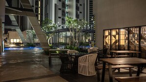 An outdoor dining area with tables and chairs, surrounded by a modern building with vertical lines and a pool in the background. - Ramada by Wyndham Meridin Johor Bahru (Iskandar Puteri)