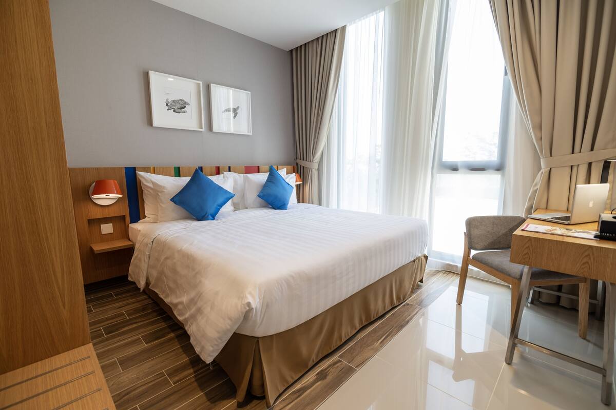 Ein Hotelzimmer mit einem großen Bett, einem Schreibtisch und einem Stuhl. An der Wand hängen zwei gerahmte Bilder, ein Nachtlicht steht auf dem Nachttisch und ein Fenster mit Vorhängen ist vorhanden.