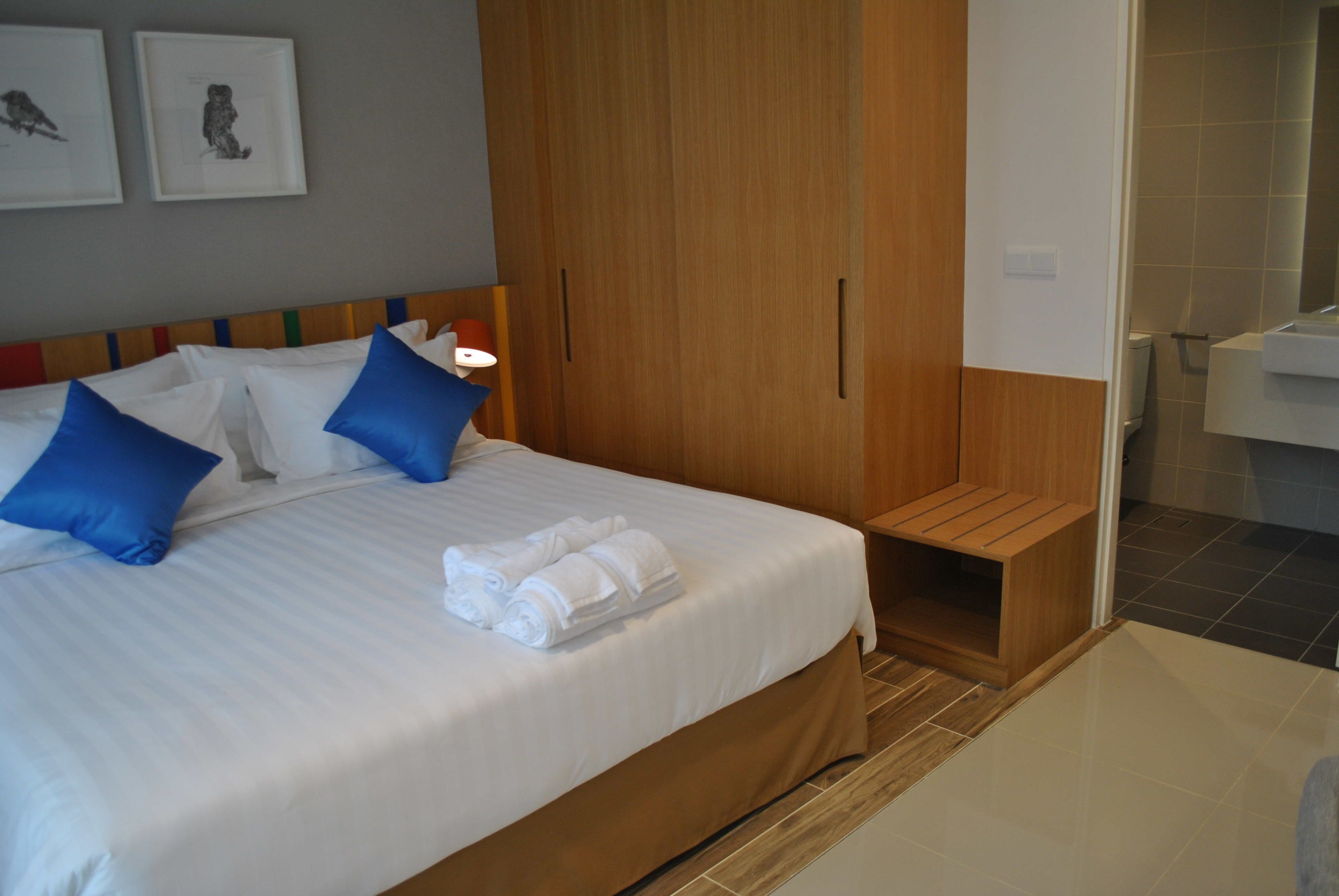 A hotel room with a large bed, blue pillows, a wooden wardrobe, a small wooden bench, and a bathroom with a white sink and tiled walls.