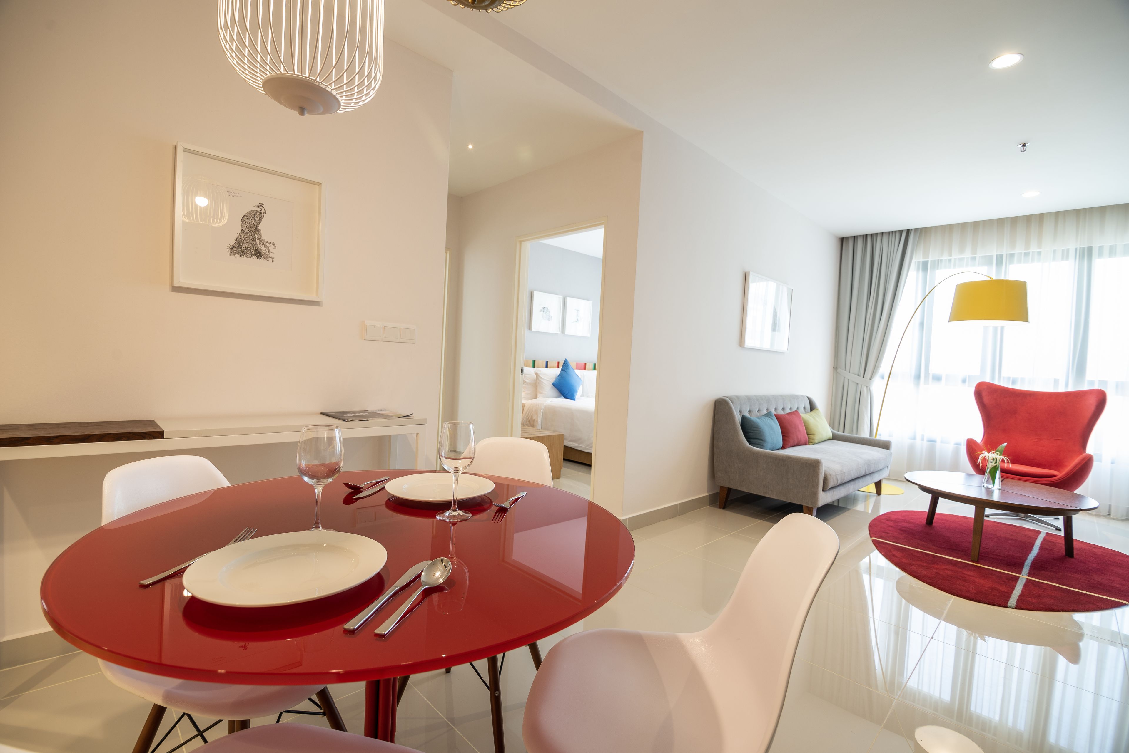 A modern hotel room with a dining area featuring a red table, white chairs, and a glass-top table. There is a gray sofa, a red armchair, and a yellow floor lamp.