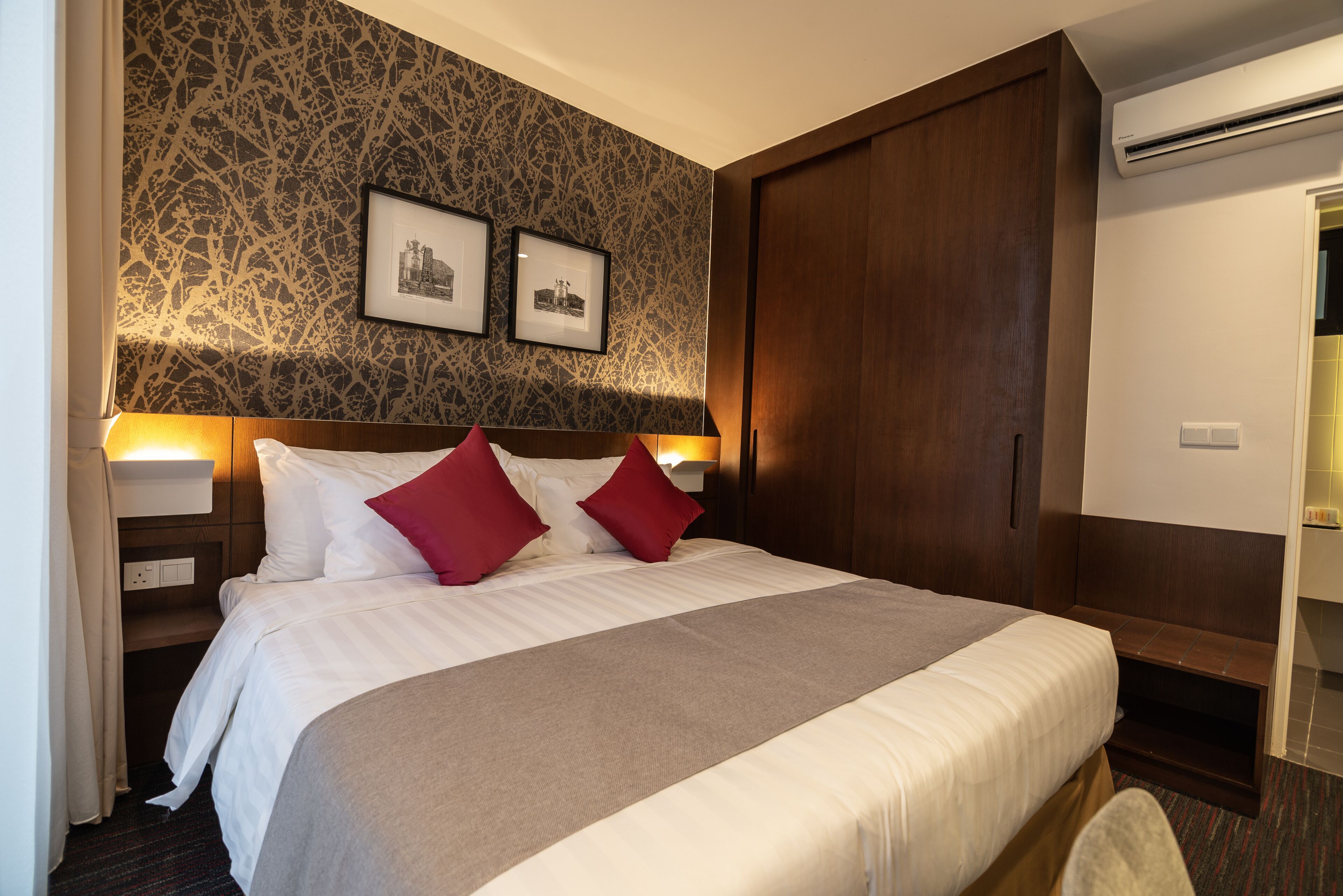 A hotel room with a large bed, two red pillows, a headboard with a picture, and a wooden wardrobe.