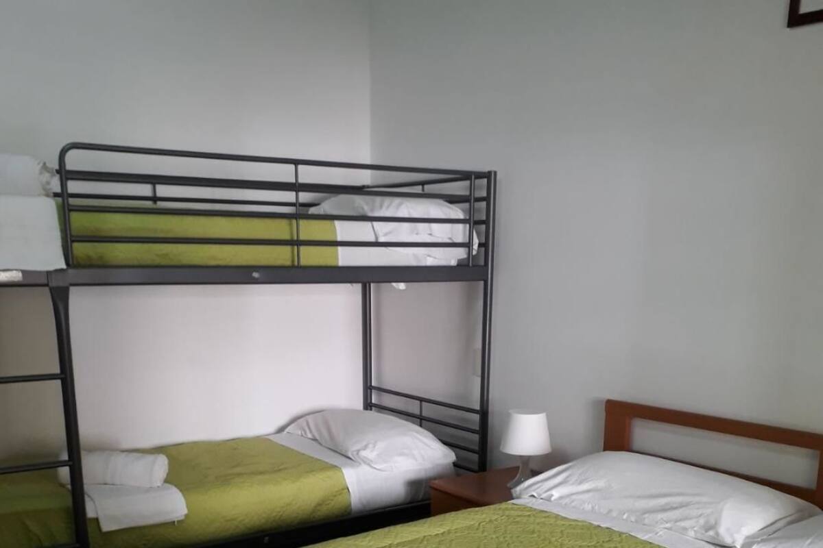 A hotel room with two bunk beds, each with green bedding and white pillows. There is a wooden nightstand with a lamp and a folded towel on each bed.
