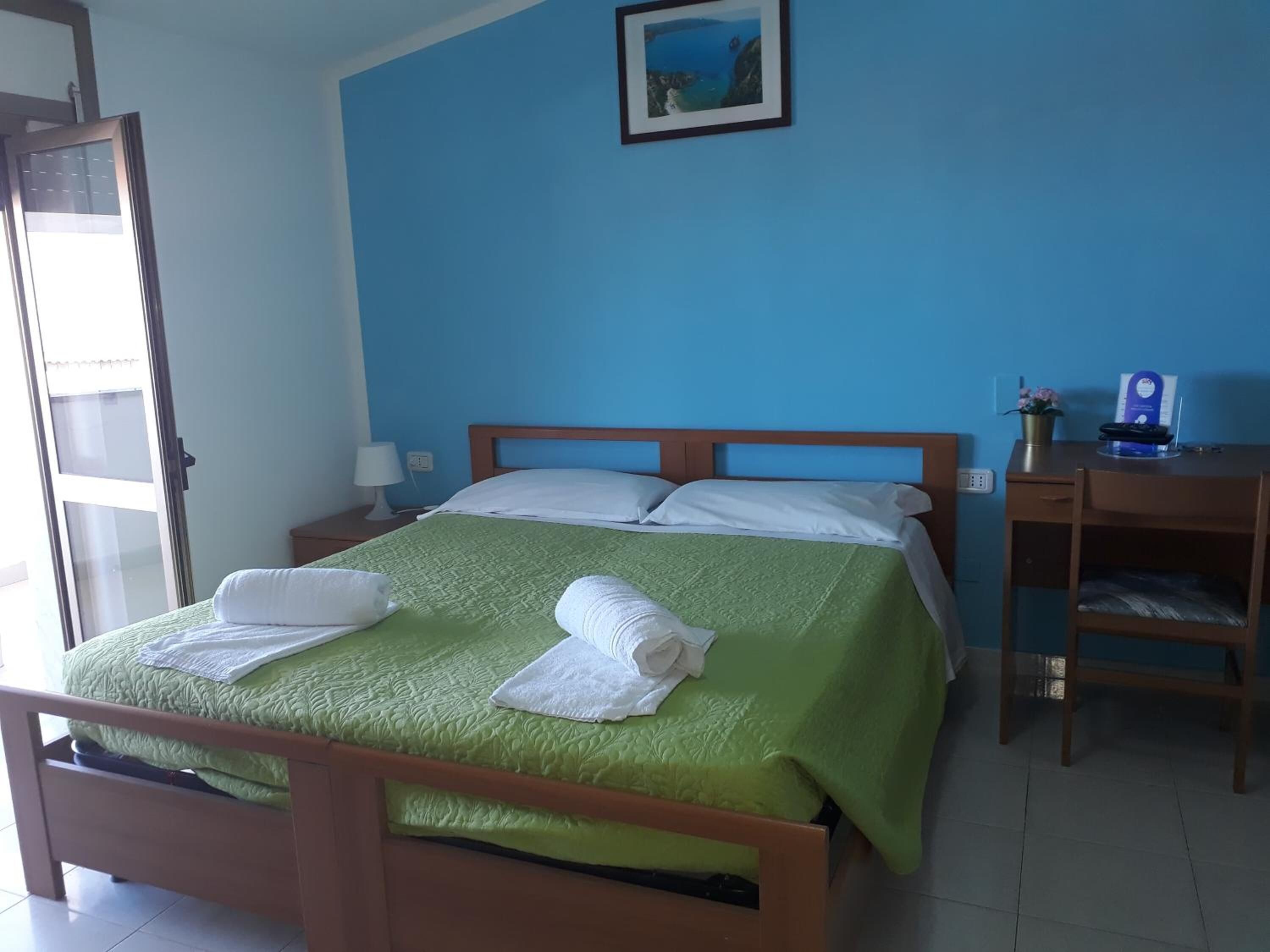A neatly made bed with green bedding and white pillows, a wooden nightstand with a lamp, a desk with a chair, and a framed picture on the wall.