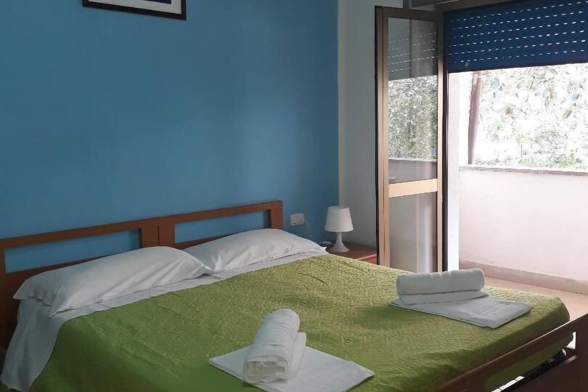 A neatly made bed with a green bedspread and white pillows, a framed picture on the wall, and a window with a view of greenery.