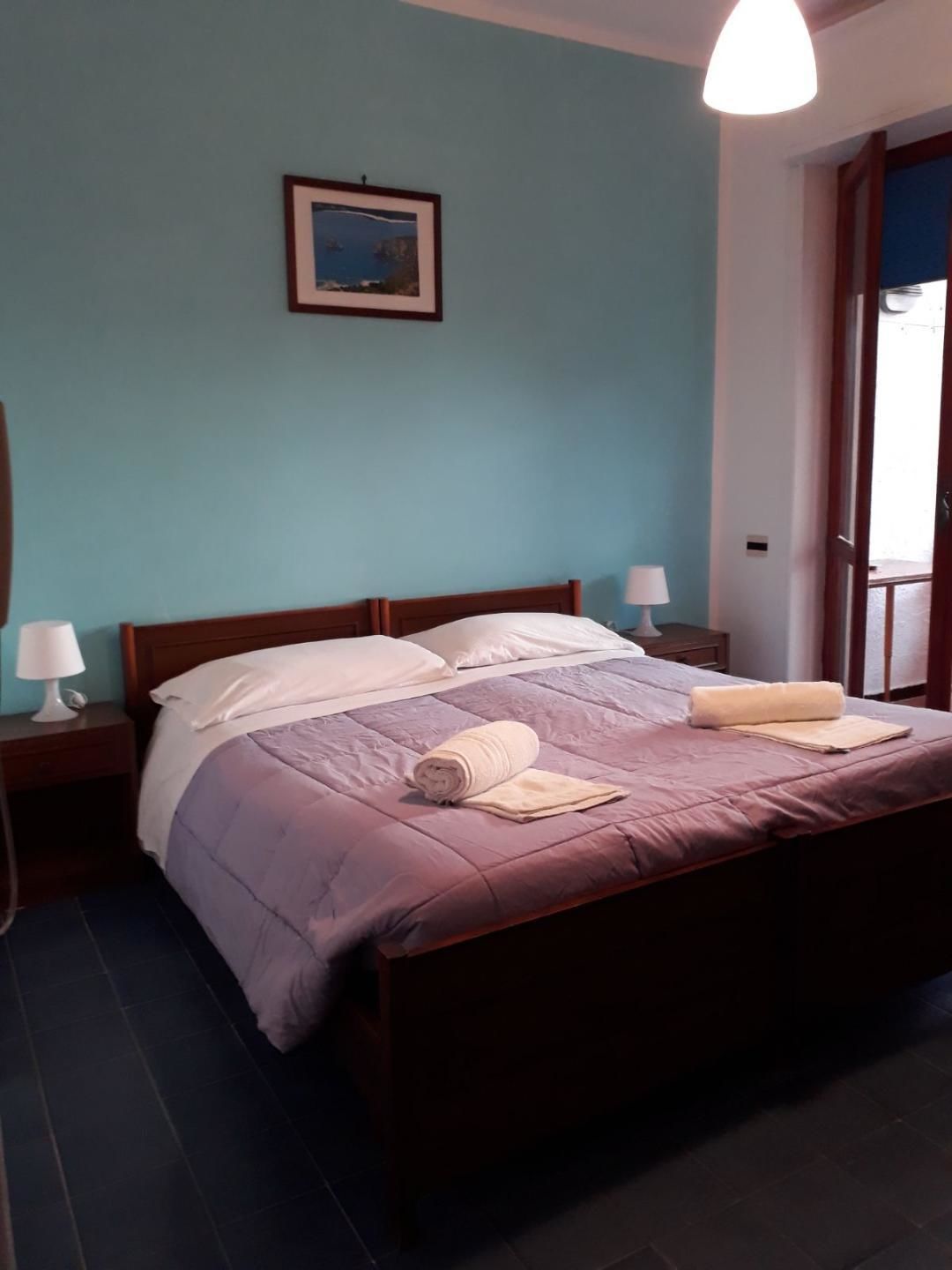 A neatly made bed with a purple bedspread and a rolled towel, a framed picture on the wall, and a bedside lamp on a wooden nightstand.