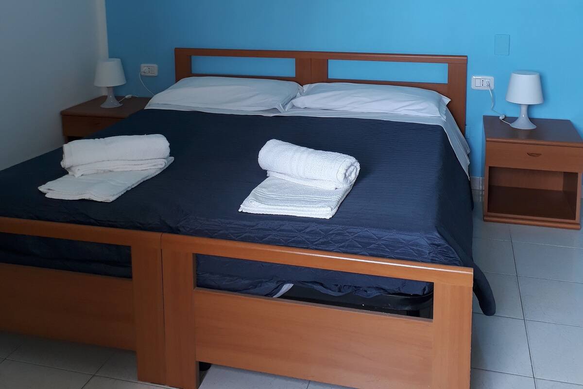 A neatly made bed with white linens and towels, a wooden headboard, and a small bedside table with a lamp.