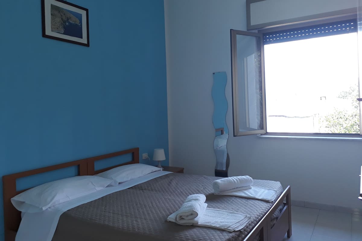 A bedroom with a bed, a window, a picture on the wall, and a bedside table with a lamp.