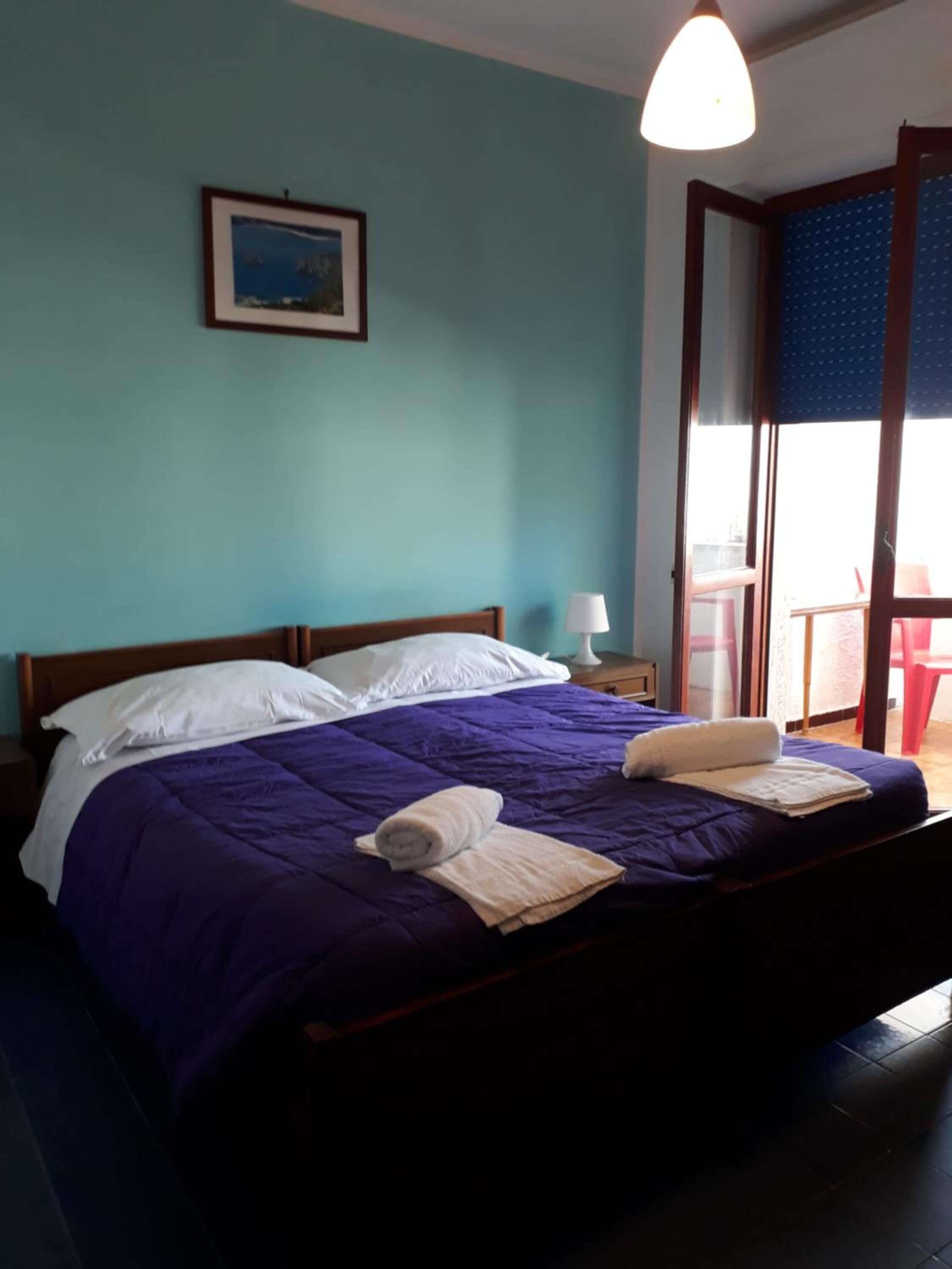 Una cama bien hecha con colcha púrpura y almohadas blancas, un cuadro en la pared y una ventana con vistas al exterior.