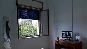 A room with a bed, a desk with a computer and a monitor, a window with a view of greenery, and a mirror on the wall. - Pensione Nettuno (Centola)