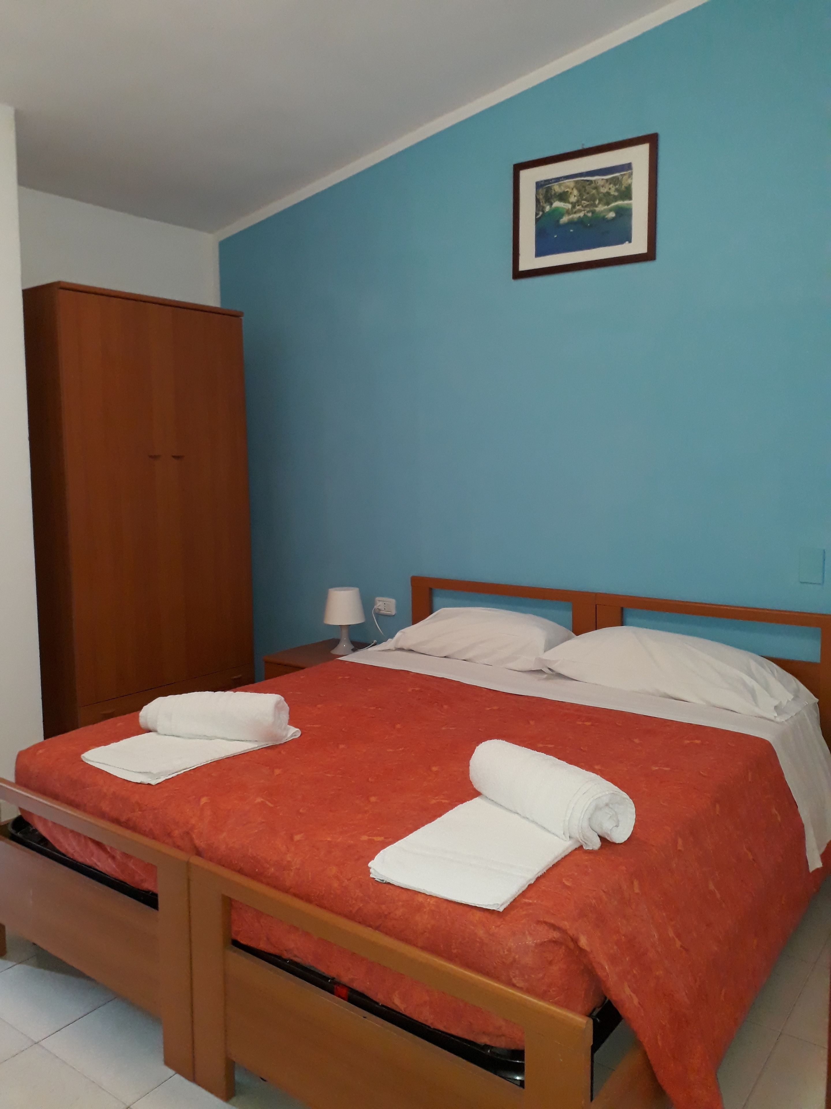 A single bed with a red bedspread, white pillows, and two rolled towels. There is a wooden wardrobe to the left and a framed picture on the blue wall above the bed.