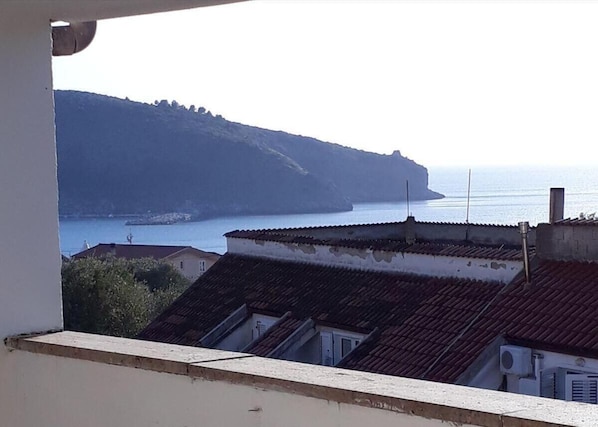 A coastal view from a balcony with terracotta-tiled rooftops and a clear view of the sea and a distant cliff.