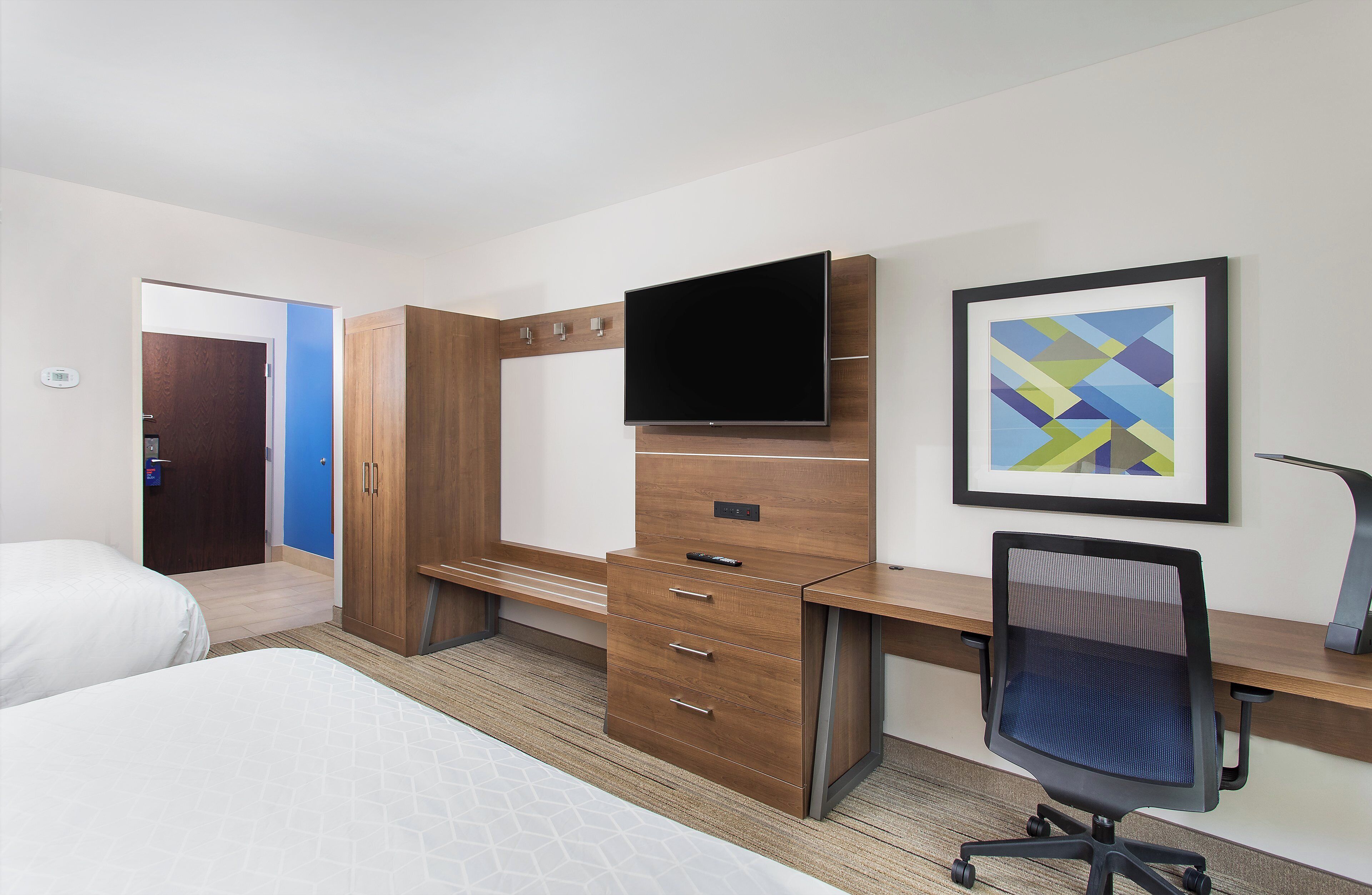 Habitación de hotel con una cama, un escritorio con televisor de pantalla plana, una silla y un cuadro abstracto enmarcado en la pared.