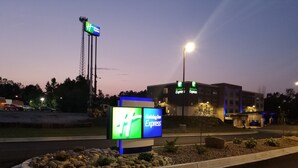 A Holiday Inn Express sign with a large 'H' logo, a parking lot, and a building with illuminated signage in the background.