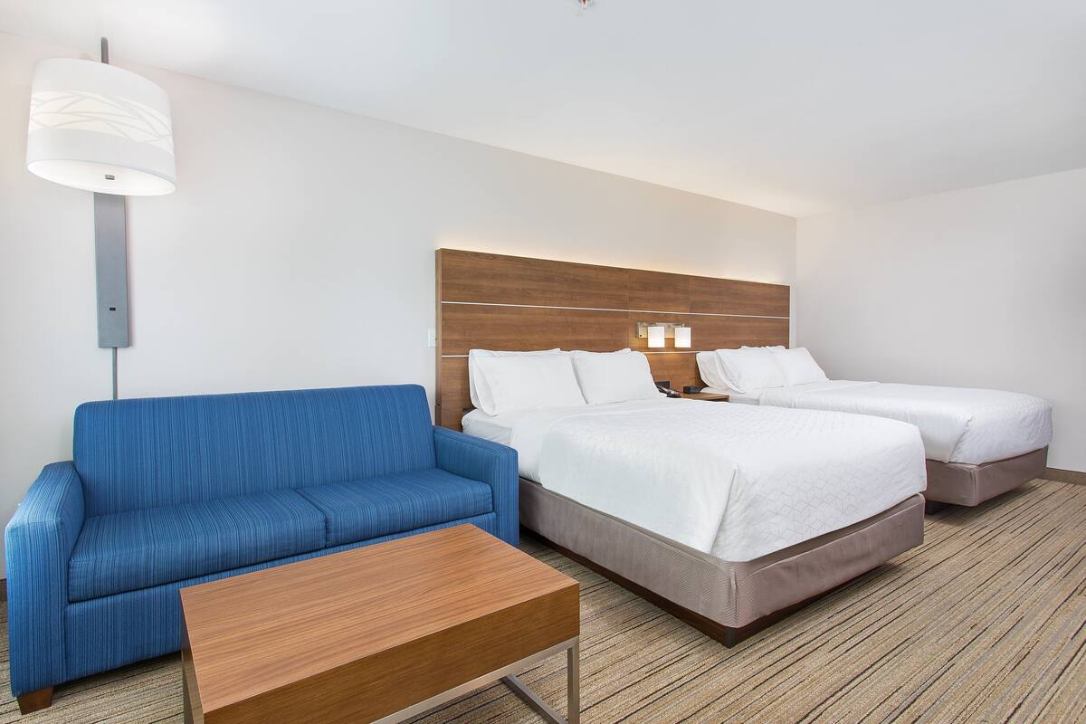 A hotel room with a blue sofa, two single beds, a wooden coffee table, and a modern light fixture.