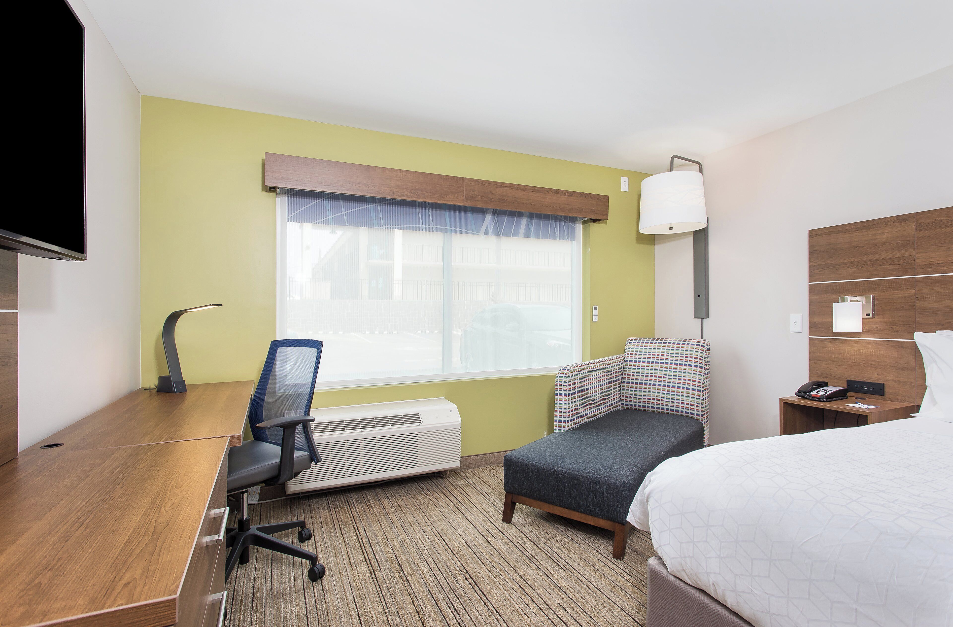 A hotel room with a bed, a desk with a chair, a standing heater, a telephone, and a window with a view of buildings.