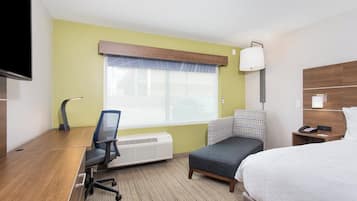 A hotel room with a bed, a desk with a chair, a standing heater, a telephone, and a window with a view of buildings.