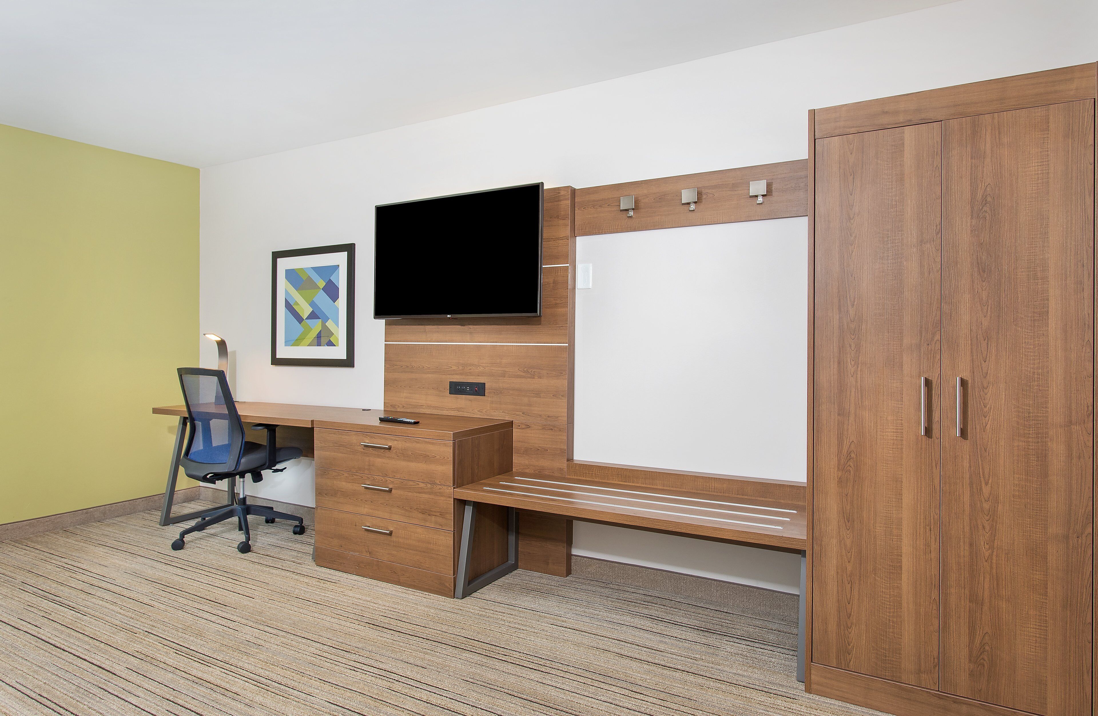 A room with a wooden desk, a chair, a television, a wardrobe, and a framed picture on the wall.