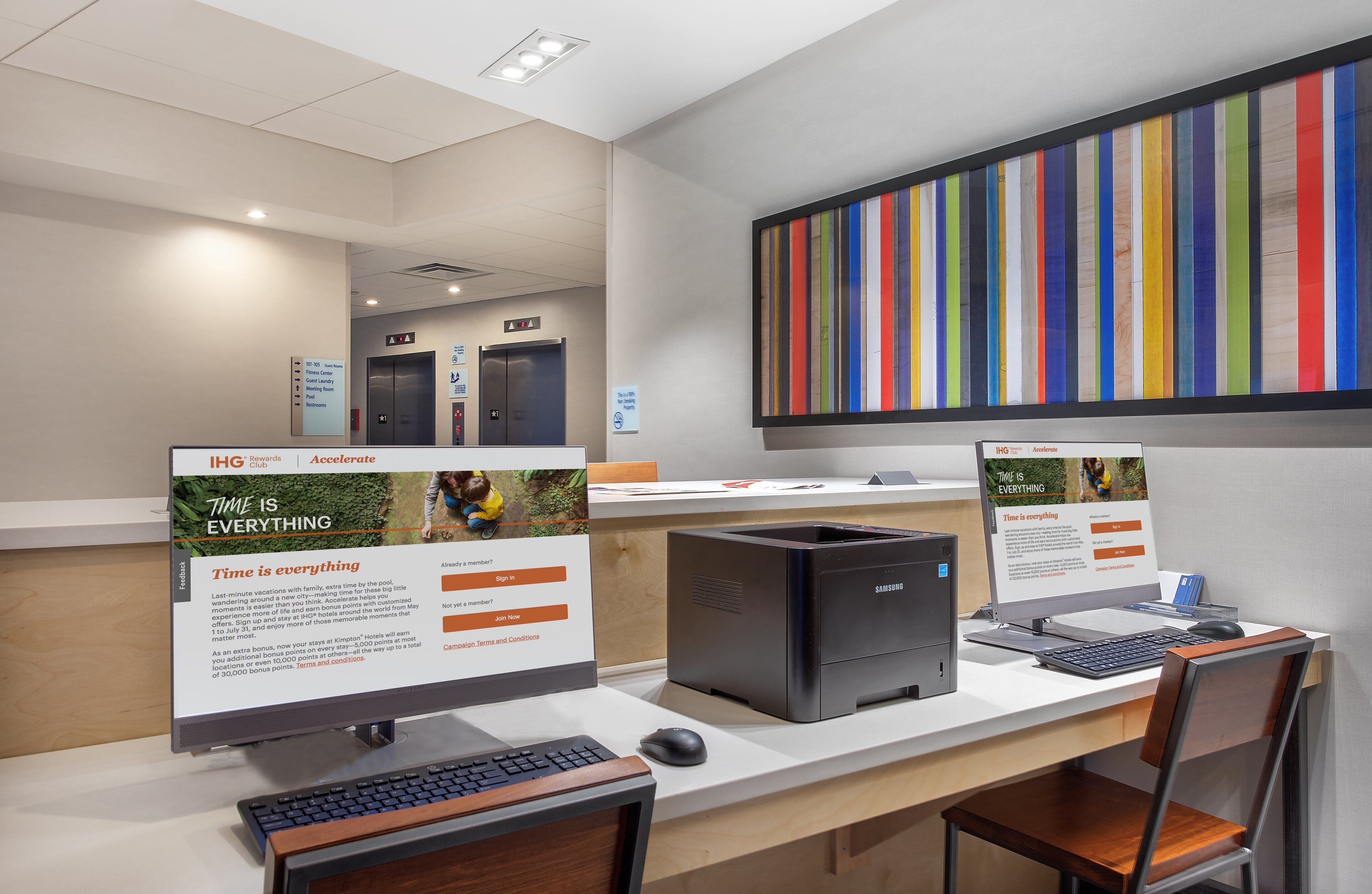 A reception desk with two computer monitors, a Samsung desktop computer, a keyboard, and a mouse. There are signs for IHG Rewards Club and Accelerate, and a colorful striped artwork on the wall.