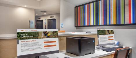 Un mostrador de recepción con dos monitores de computadora, una computadora de escritorio Samsung, un teclado y un ratón. Se observan carteles de IHG Rewards Club y Accelerate, además de una obra de arte colorida con rayas en la pared.