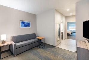A hotel room with a grey sofa, a small wooden side table with a lamp, a framed abstract painting on the wall, and a television on a wooden stand.