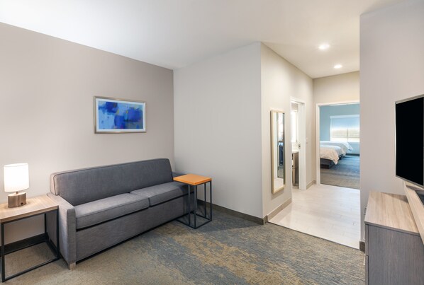 A hotel room with a grey sofa, a small wooden side table with a lamp, a framed abstract painting on the wall, and a television on a wooden stand. - Holiday Inn Lubbock South by IHG (Lubbock)