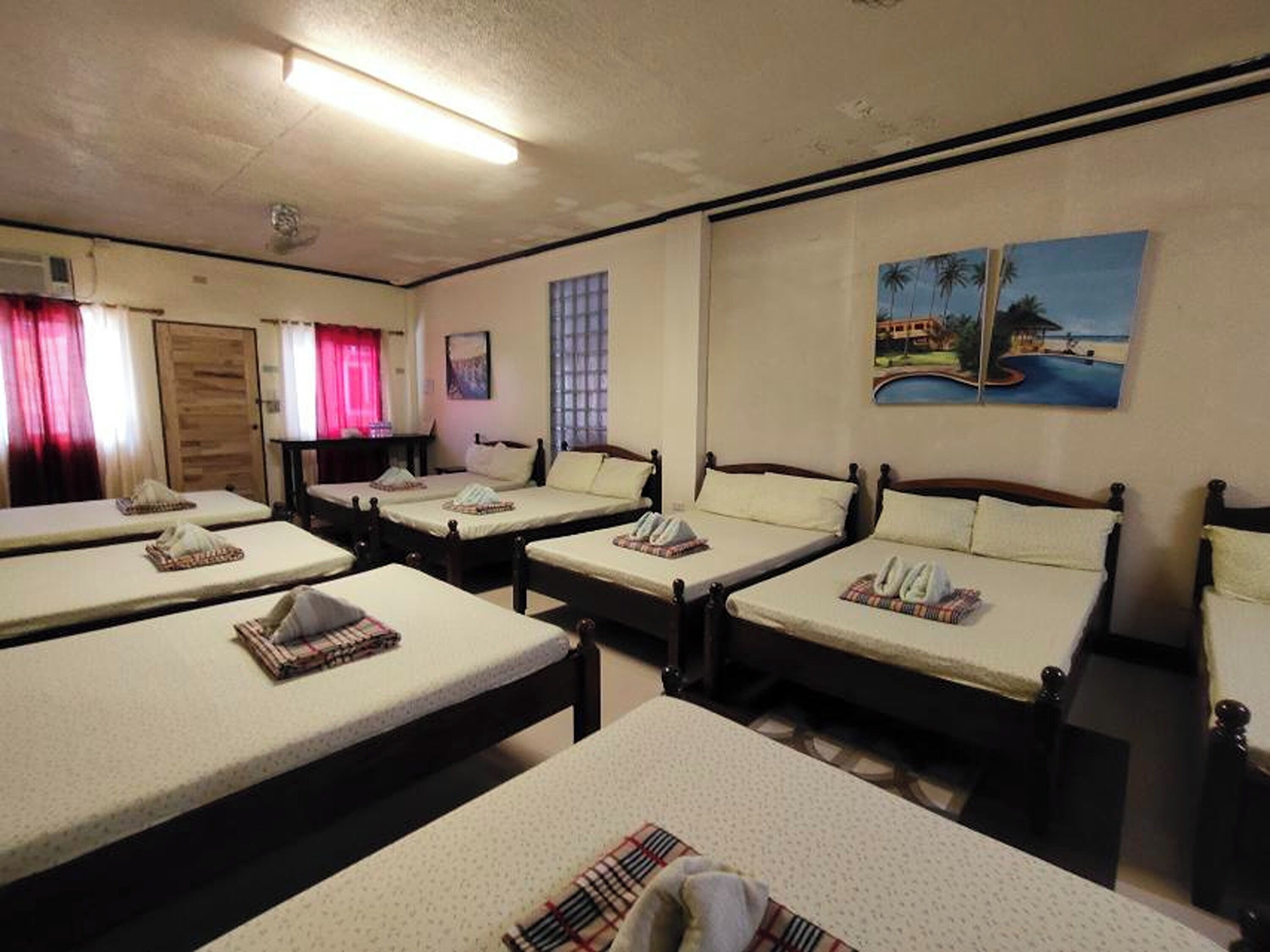 Une chambre d’hôtel avec quatre lits simples, une porte en bois, une fenêtre avec des rideaux, un tableau représentant une scène de plage et un ventilateur de plafond.