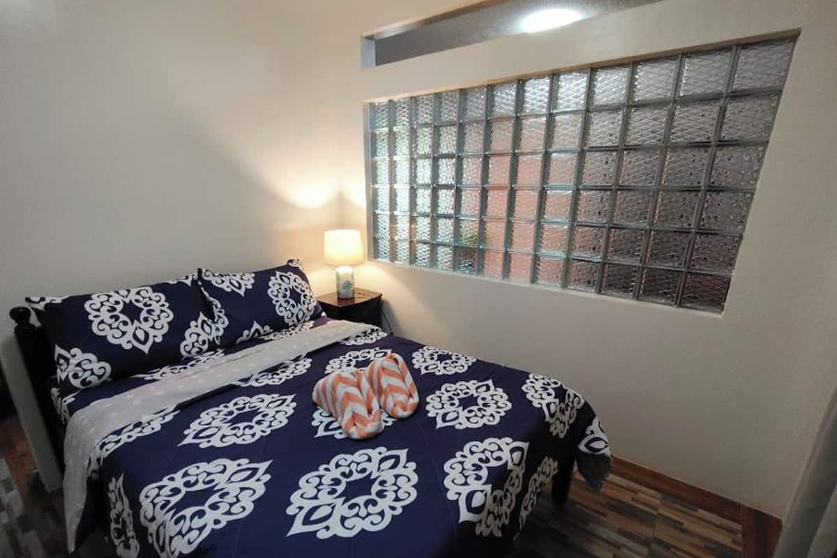 A bedroom with a bed, a nightstand, a lamp, and a window with a view of the outside.