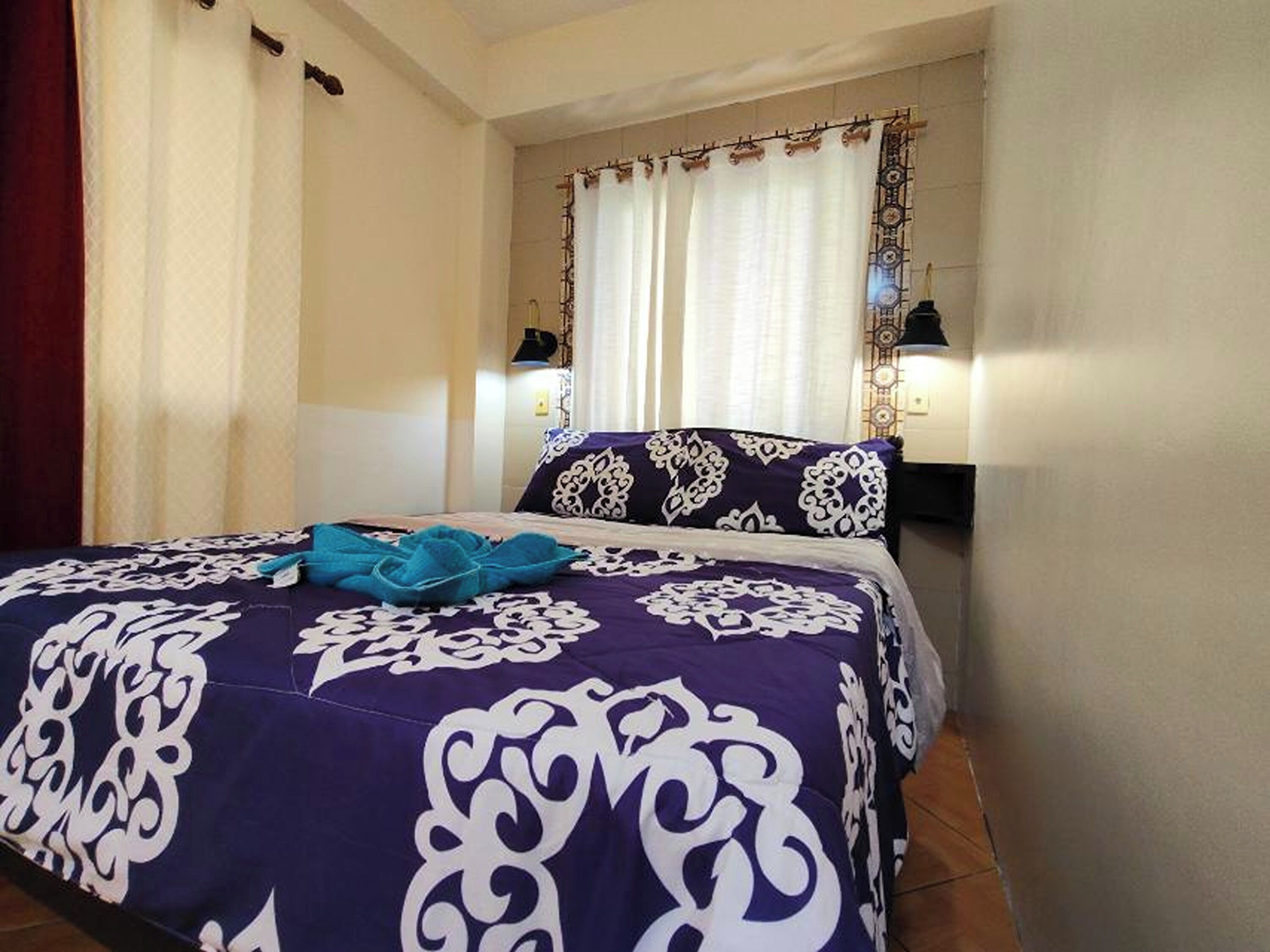 Een net opgemaakt bed met een bedspread met patroon, een blauwe plaids en een decoratief kussen. Er is een raam met gordijnen en een wandlamp.