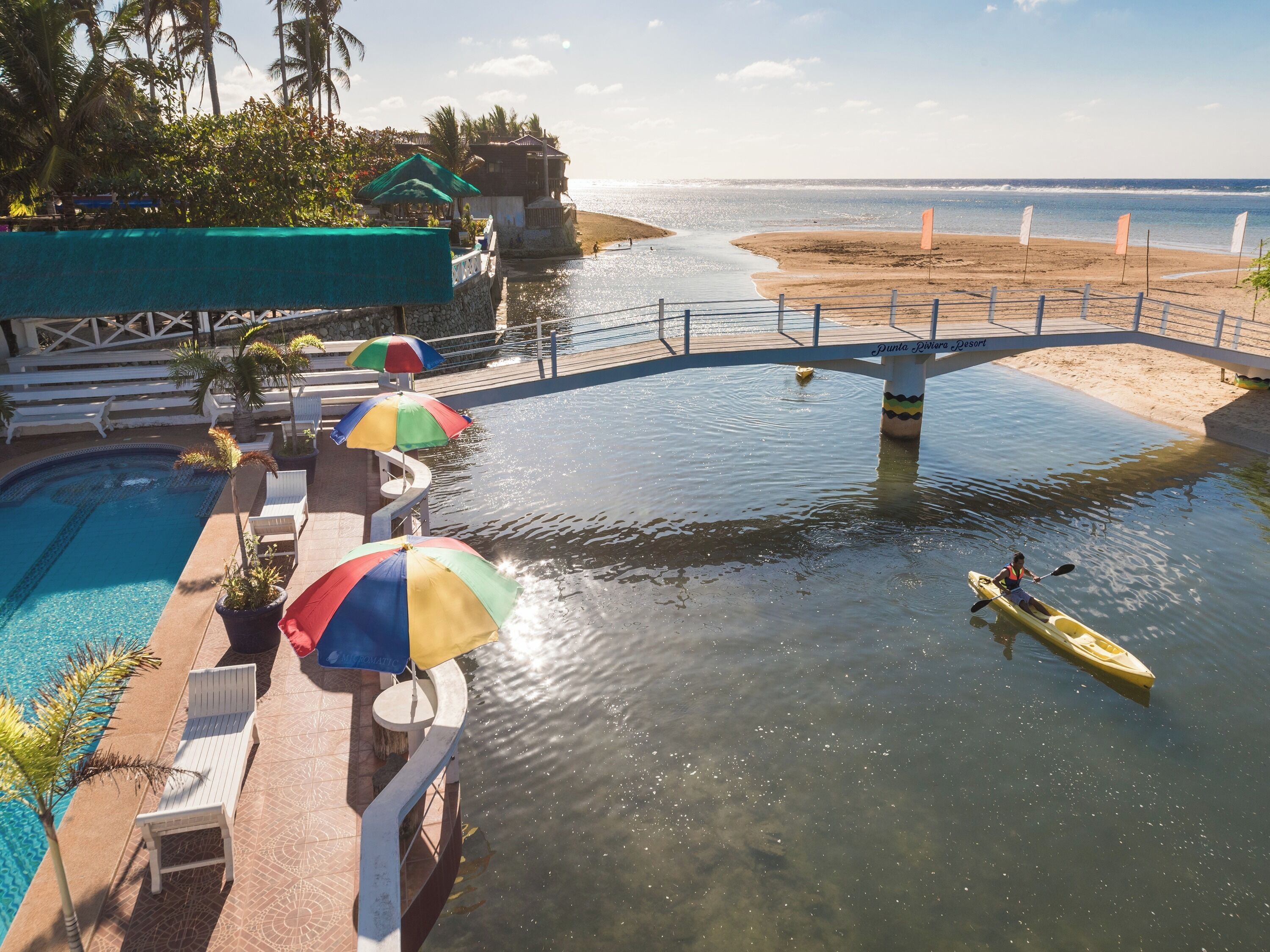 A pool area with lounge chairs and umbrellas, a bridge over water, and a kayaker.