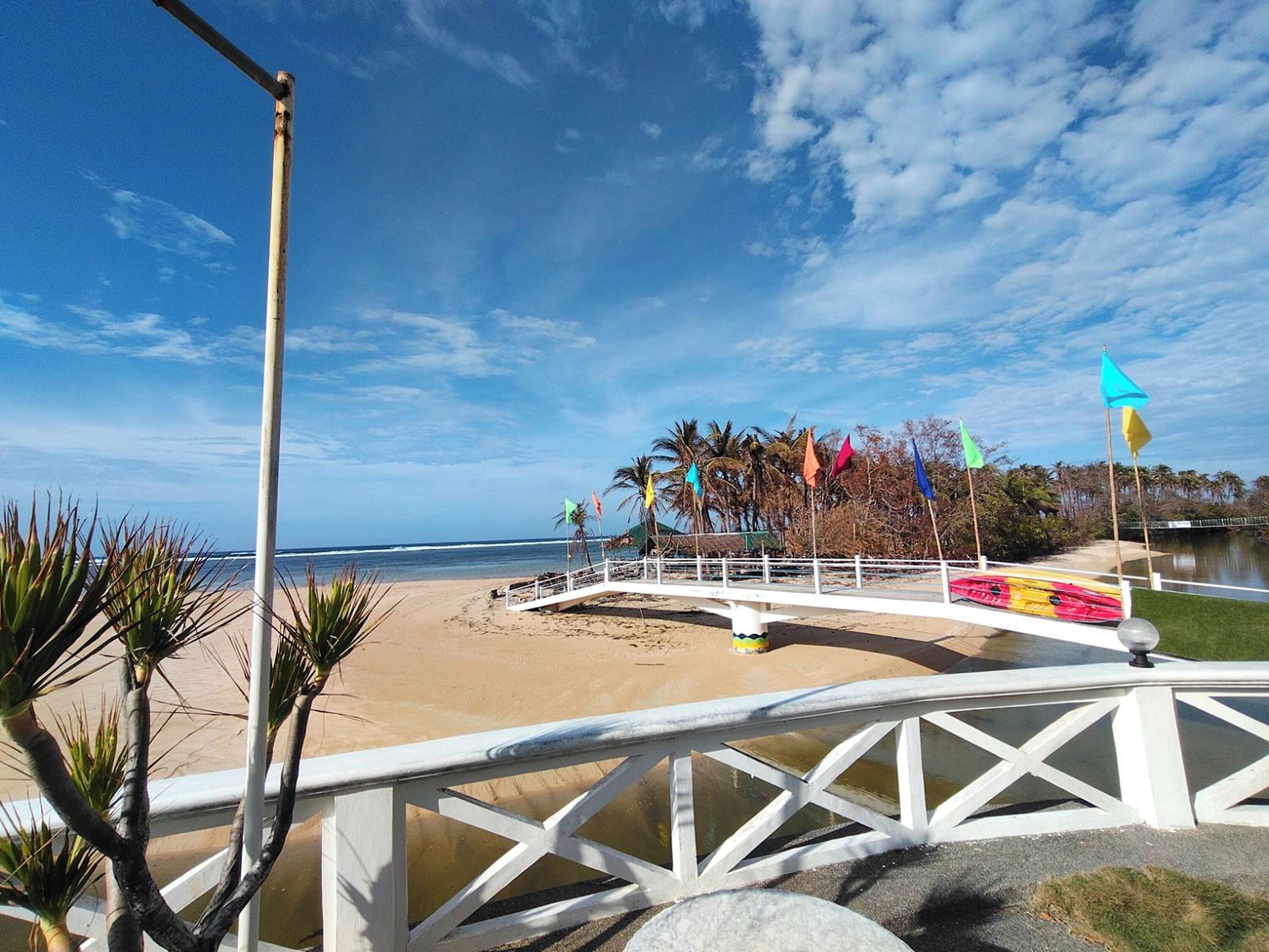 A beach with a white bridge, palm trees, and colorful flags.