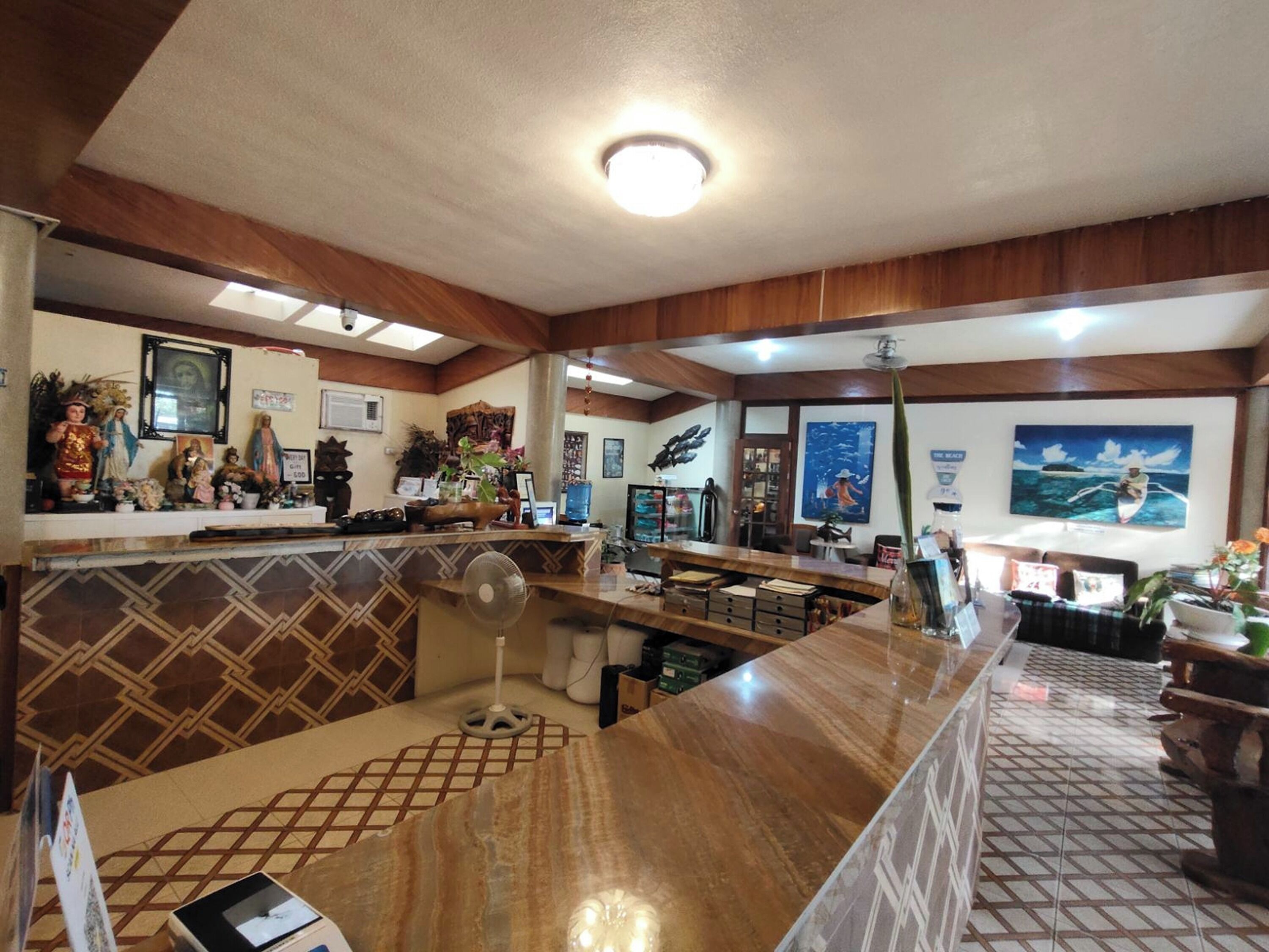 A hotel lobby with a marble reception desk, a fan, and various decorations including religious statues and artwork.