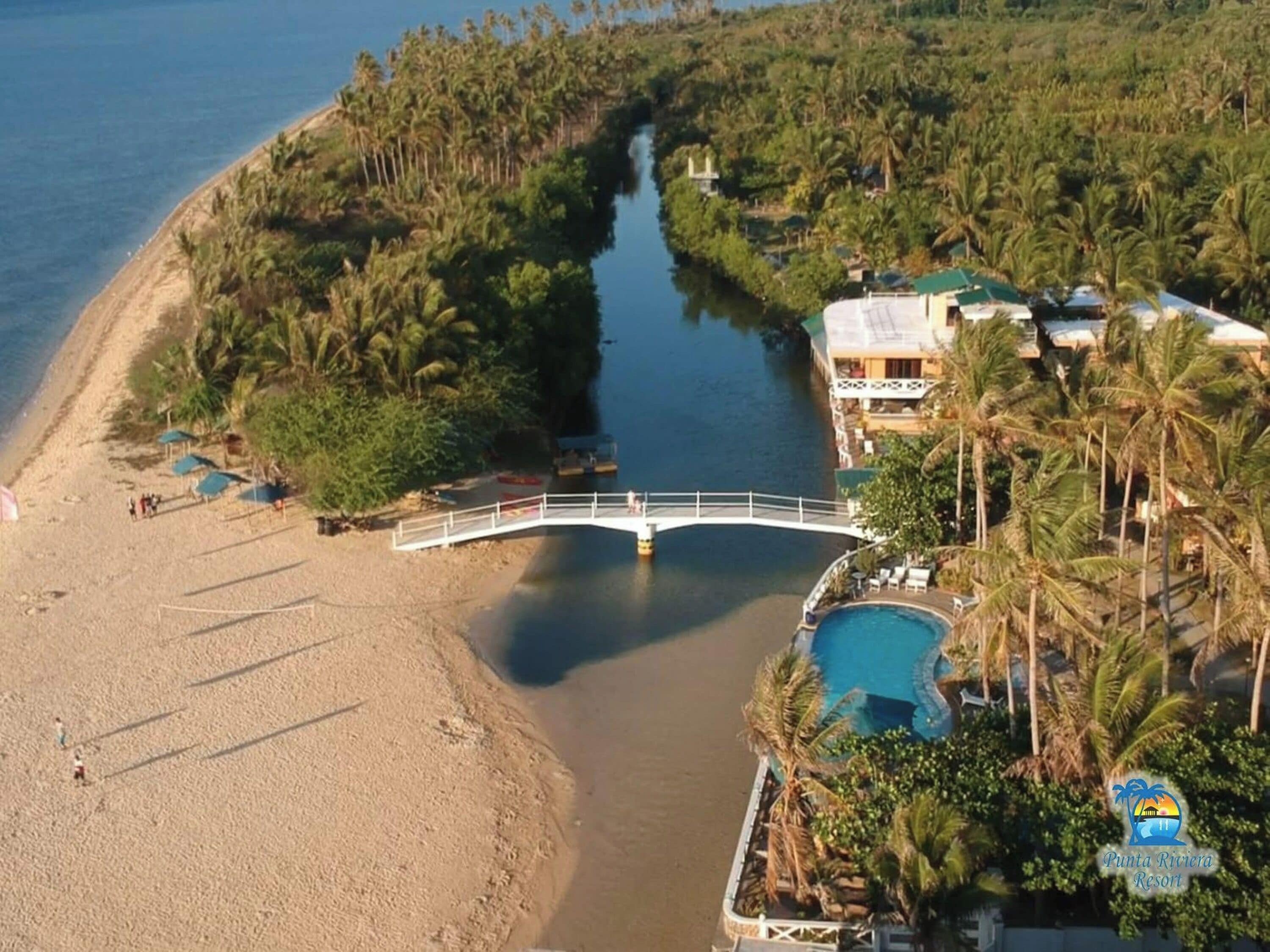 Vue aérienne d’un complexe hôtelier avec un pont enjambant un canal, une plage et un espace piscine entouré de palmiers.