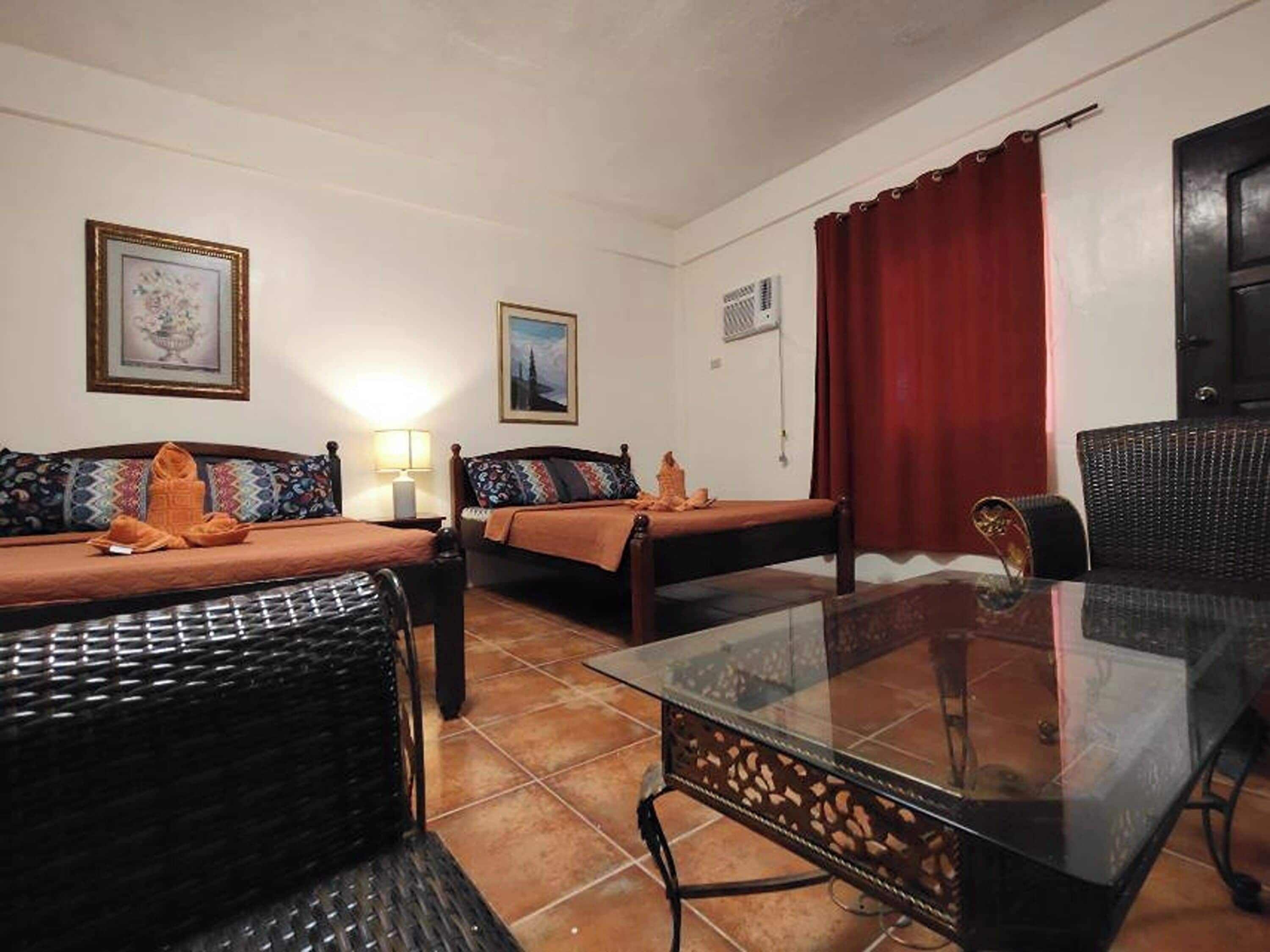 A room with two beds, a glass table, wicker furniture, and a red curtain.