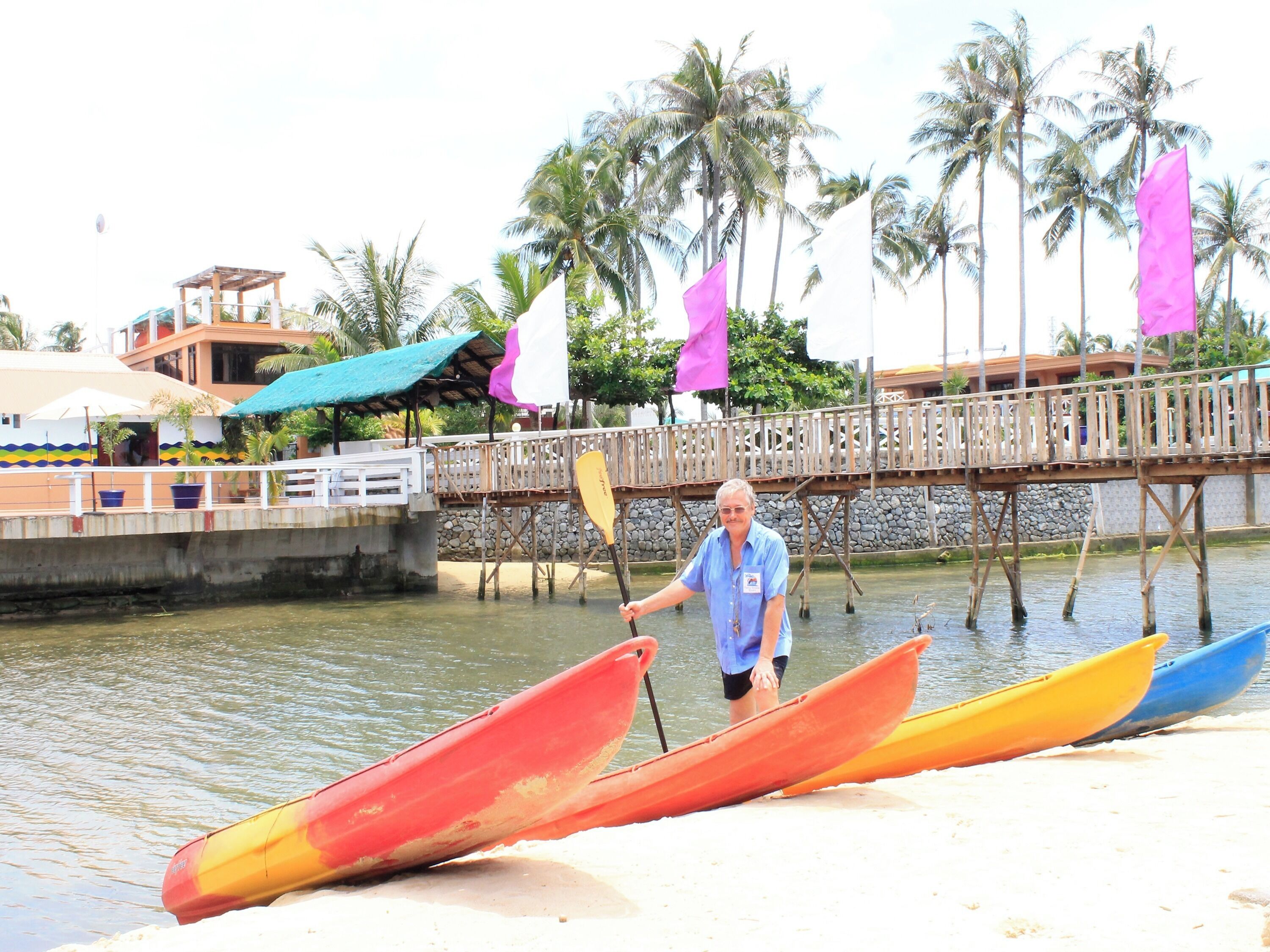 Un uomo in piedi vicino a un molo, con canoe, un ponte di legno e palme sullo sfondo.