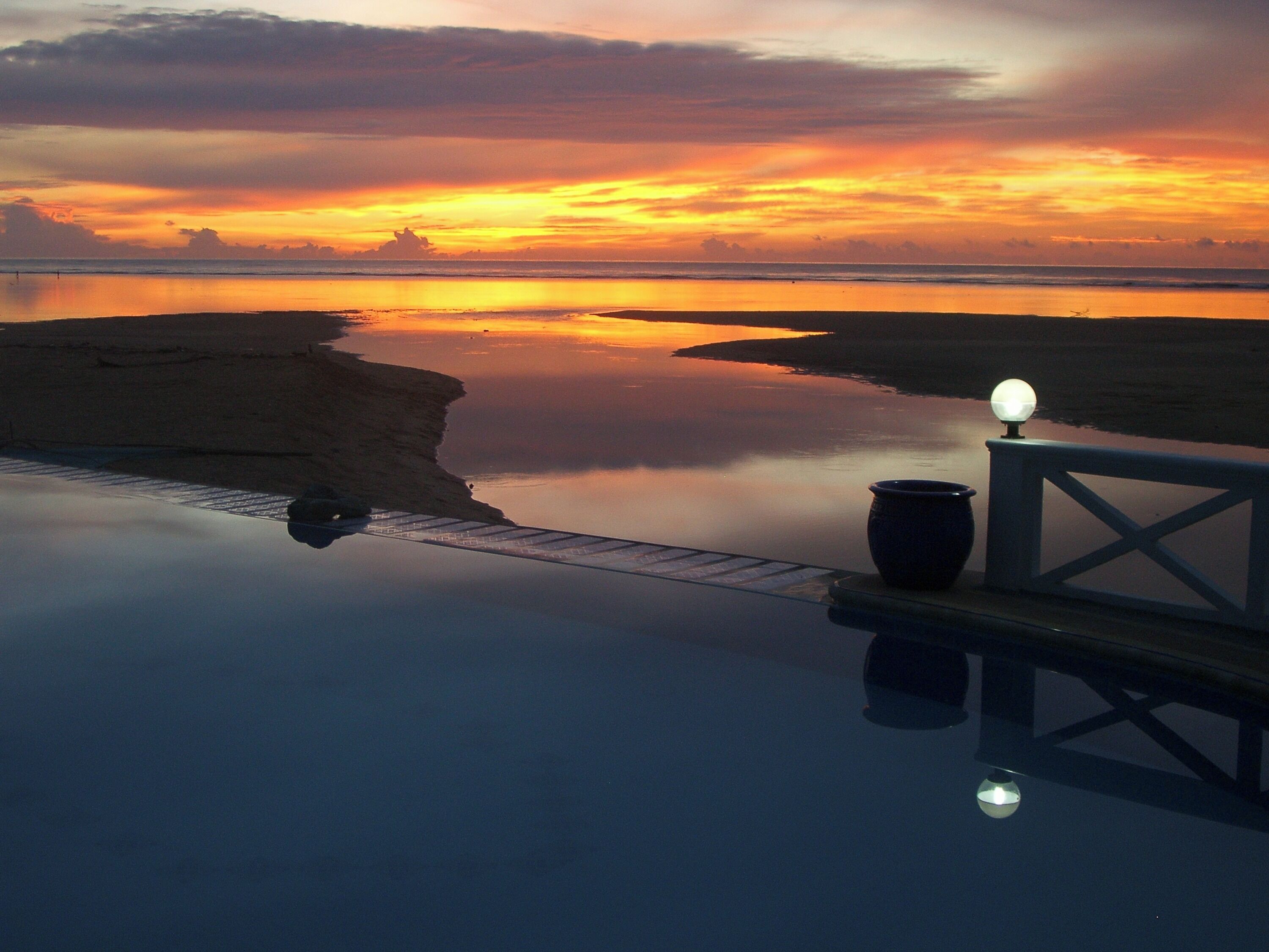 Una vista serena del atardecer desde un balcón con una piscina tranquila, una lámpara y una planta en maceta.