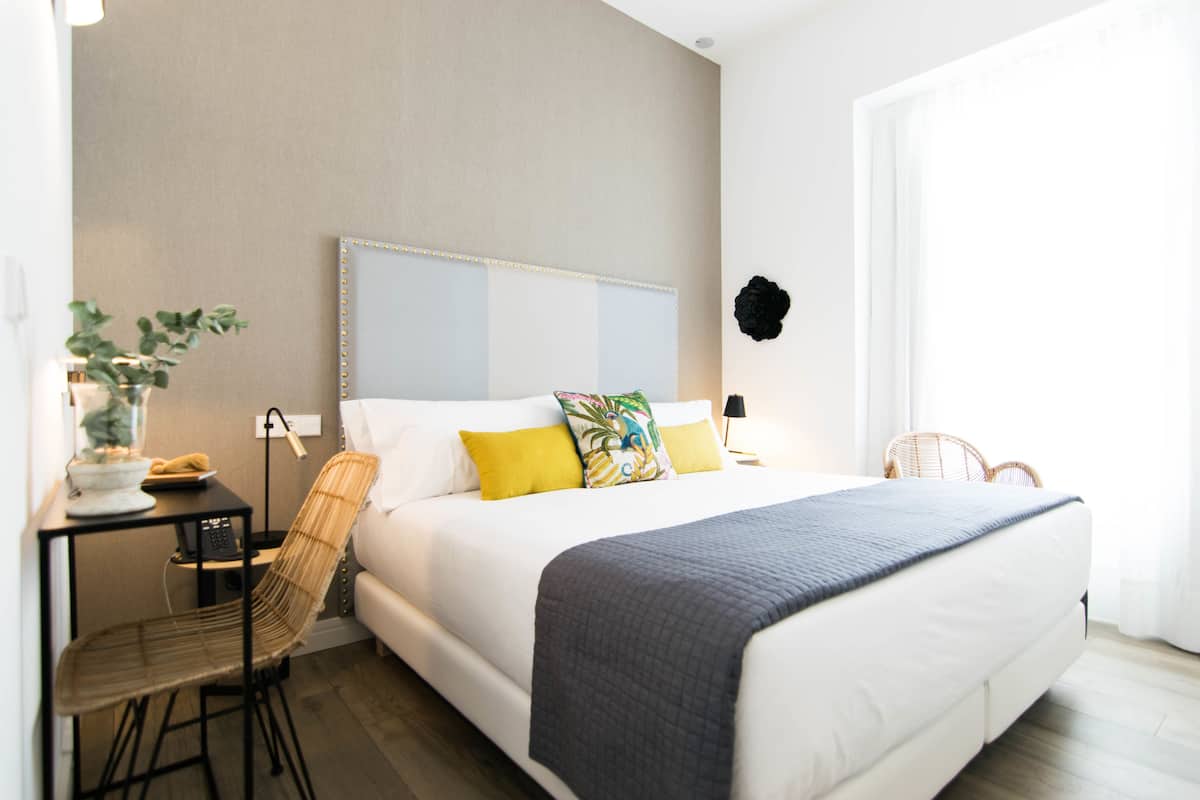 A hotel room with a bed, a desk with a chair, a potted plant, and a wall-mounted decorative item.