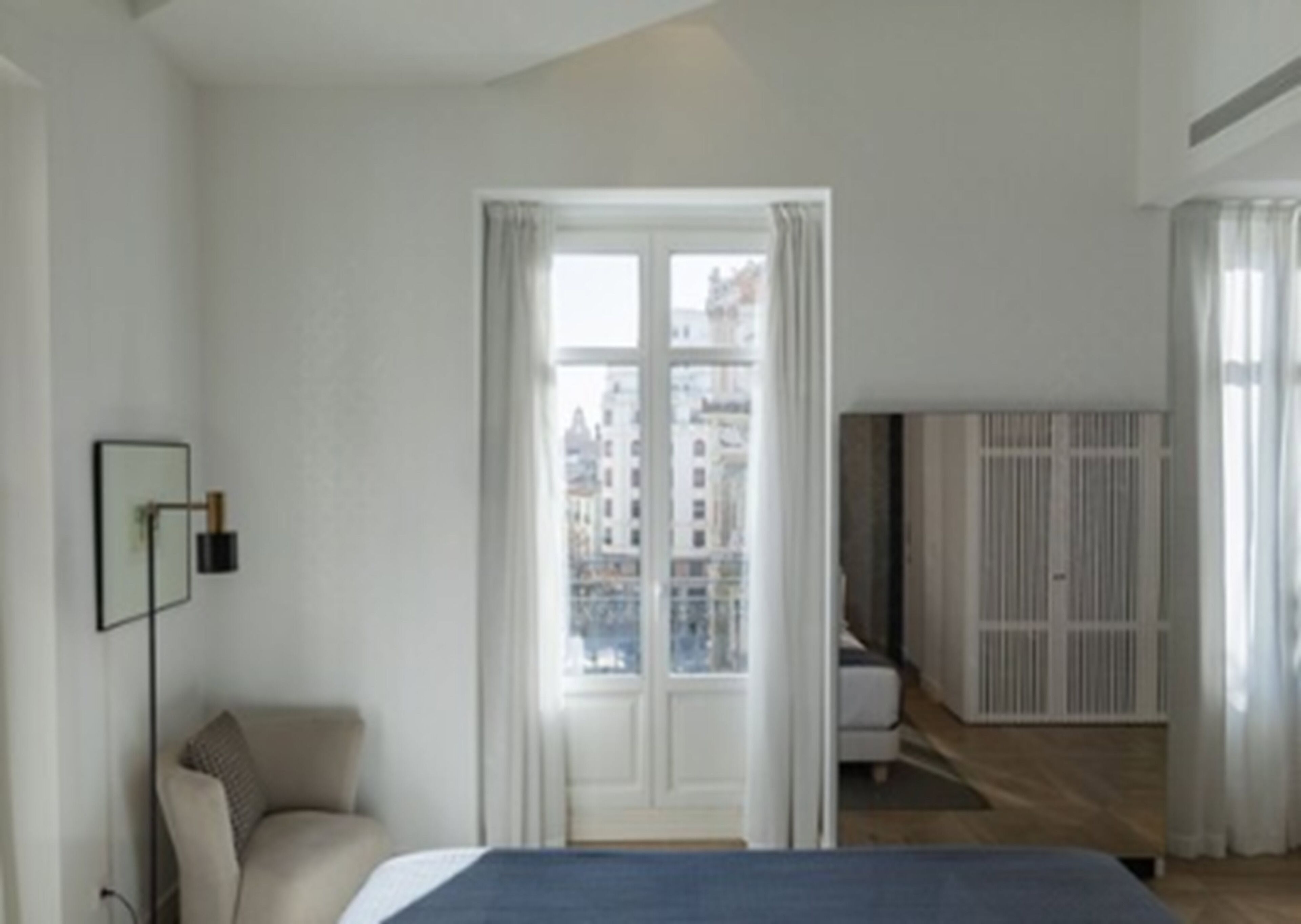 A modern hotel room with a large bed, a sofa, a view of the city through a window, and a wardrobe.
