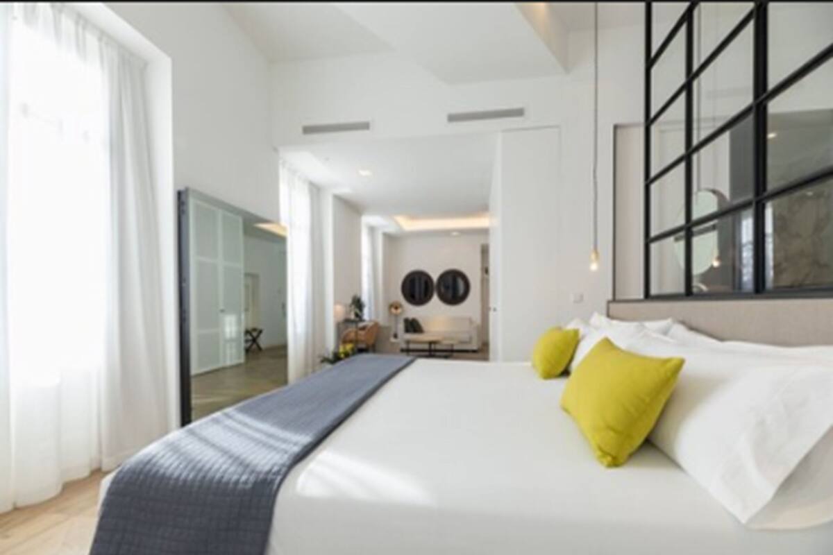 A modern hotel room with a large bed, a view of the city, and a dining area in the background.