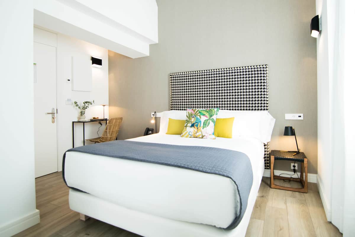 A modern hotel room with a large bed, a bedside table with a lamp, a small side table, and a wall-mounted light fixture.