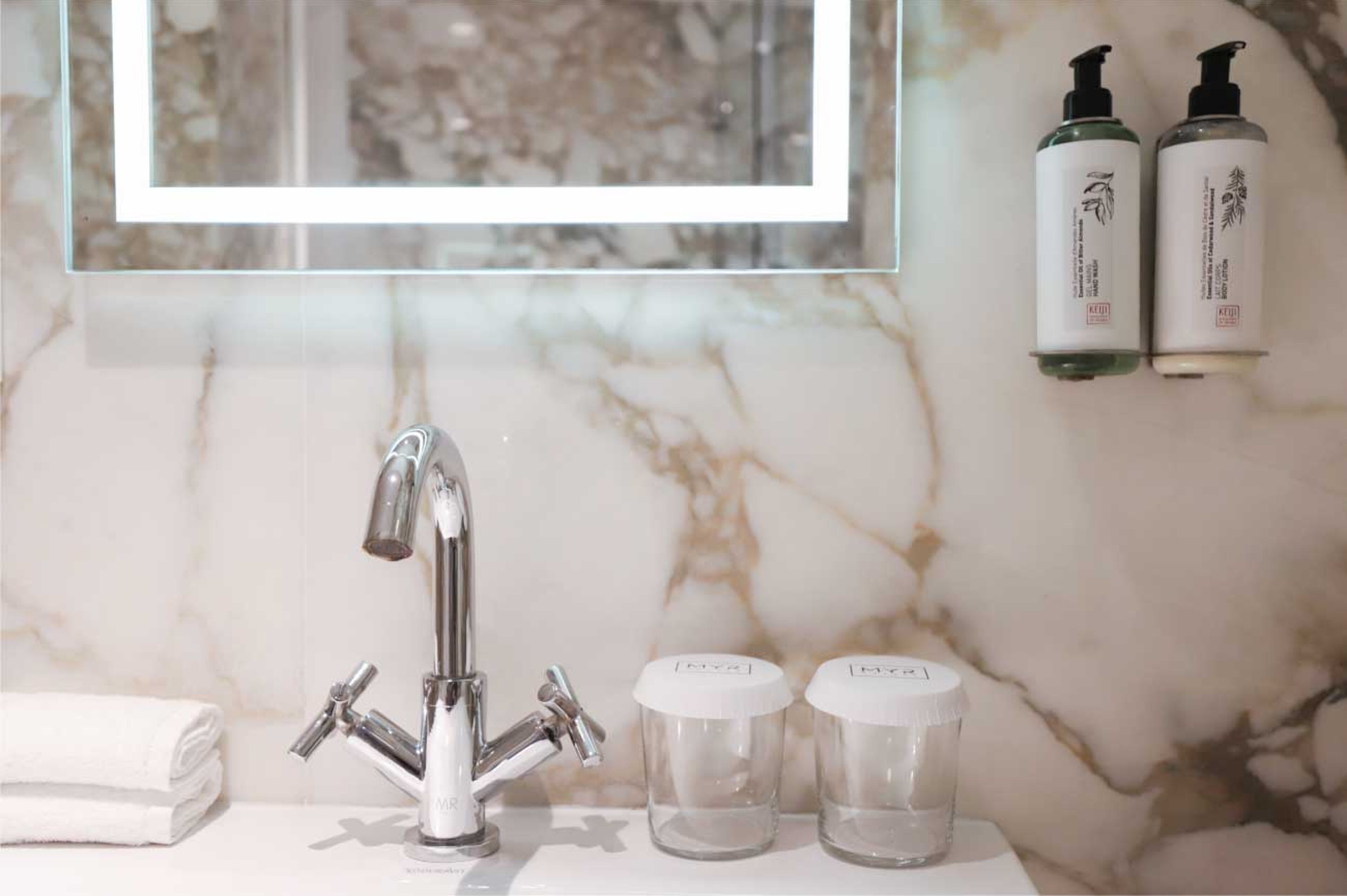Un baño con lavamanos de mármol, una llave moderna y dos botellas de productos para el cuidado de la piel en la pared.