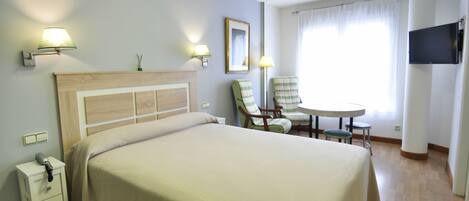 Quarto de hotel com cama, mesa de cabeceira, luminária, cadeira, mesa e televisão.