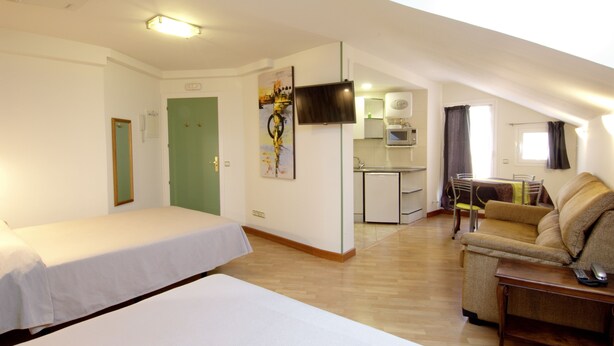 Una habitación con cama, sofá, mesa de comedor con sillas, zona de cocina y televisor montado en la pared.