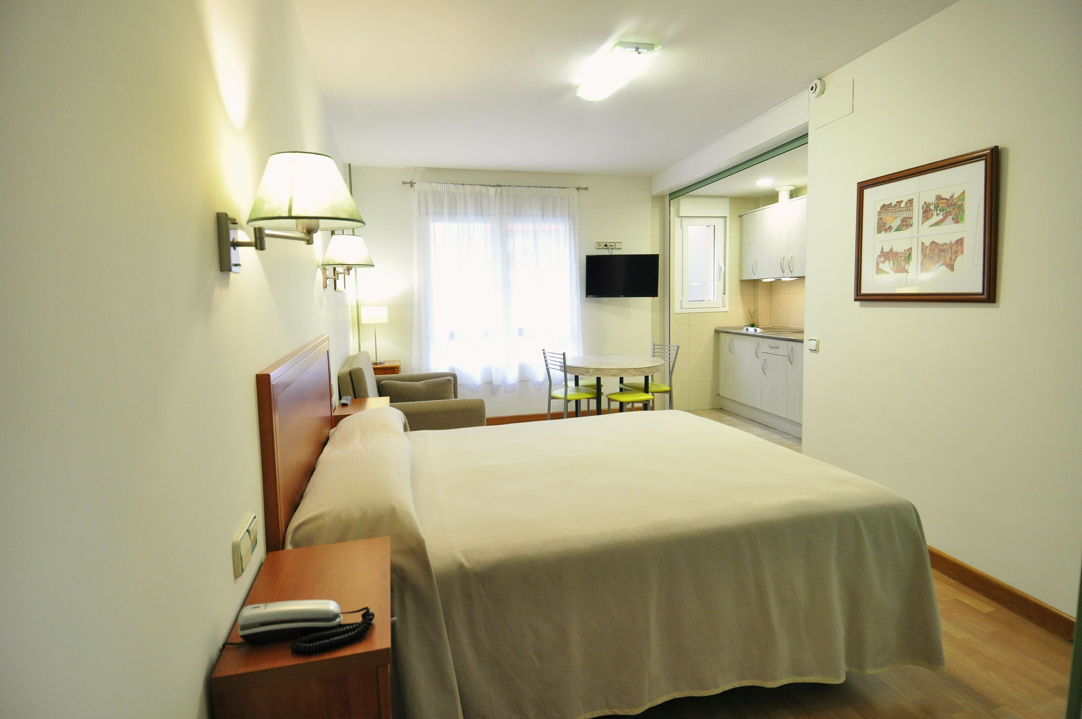 A hotel room with a bed, bedside table, lamp, chair, desk, and a kitchenette area.
