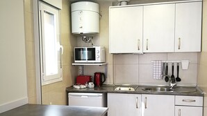 Cocina compacta con armarios blancos, microondas, cafetera y fregadero. Hay dos taburetes junto a una mesa pequeña.