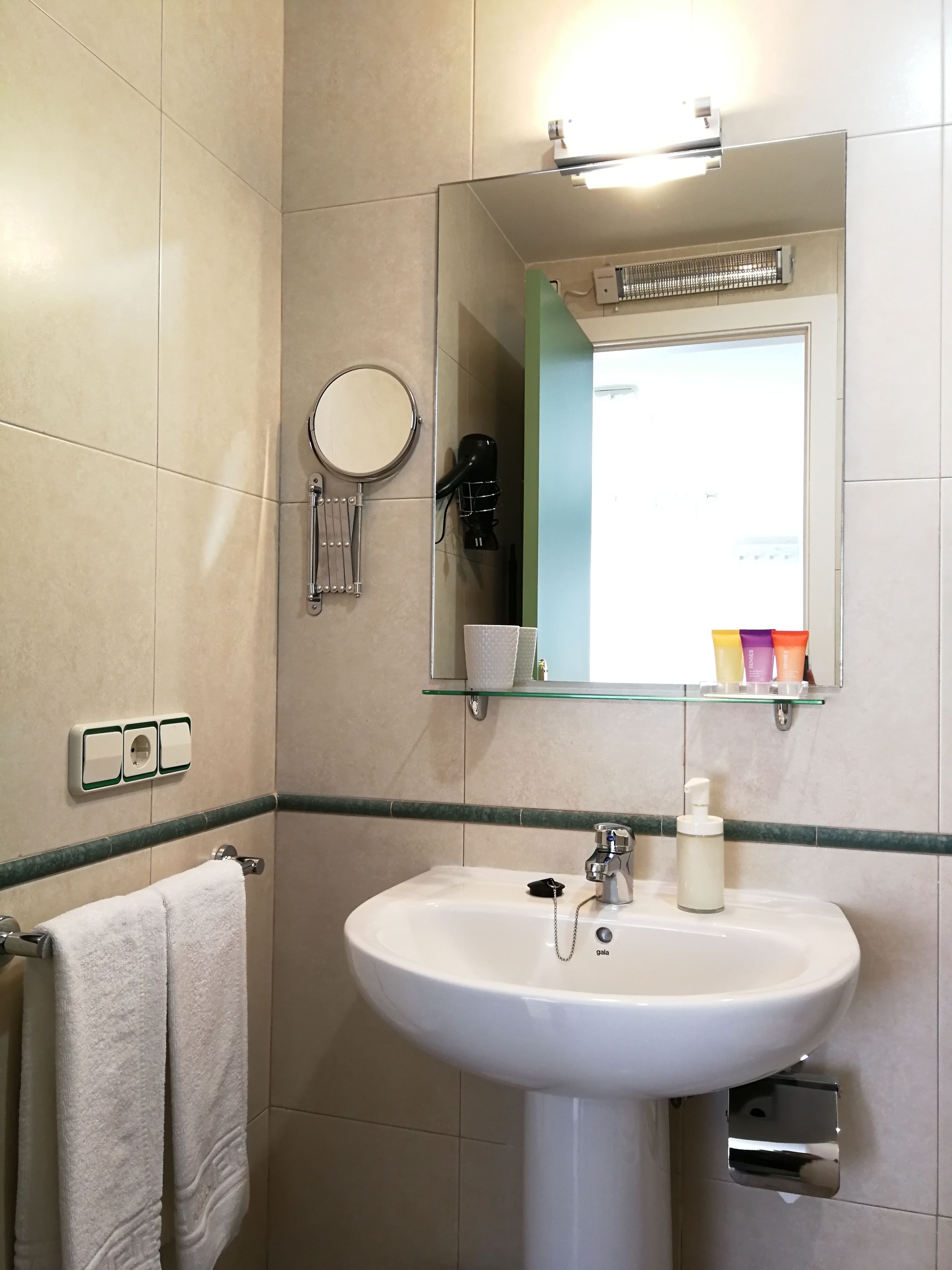 Un bagno con un lavandino bianco, uno specchio, un portascopino e uno scaffale con articoli da toilette.