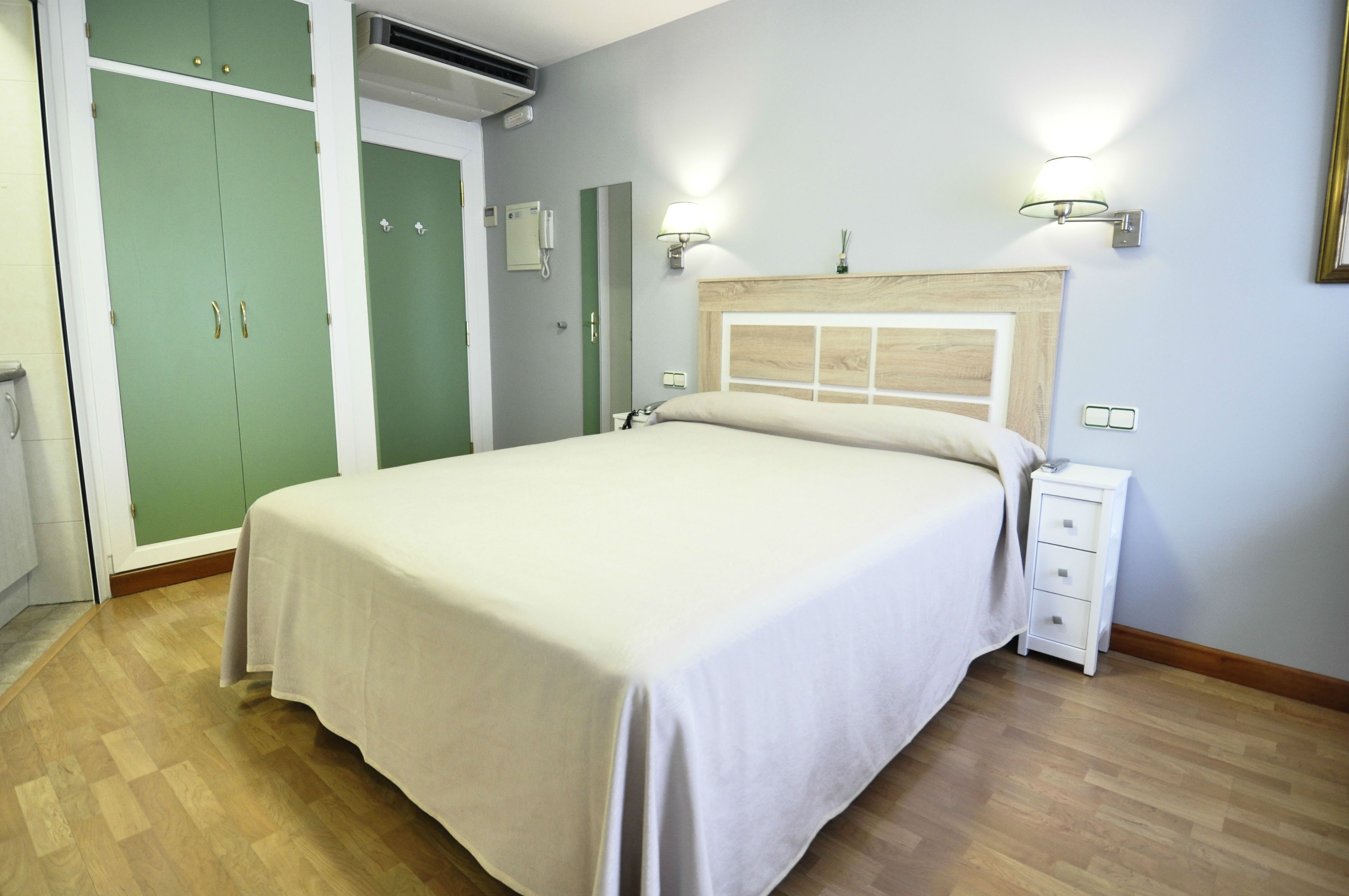 A bedroom with a bed, bedside table, nightstand, wardrobe, and a wall-mounted lamp.